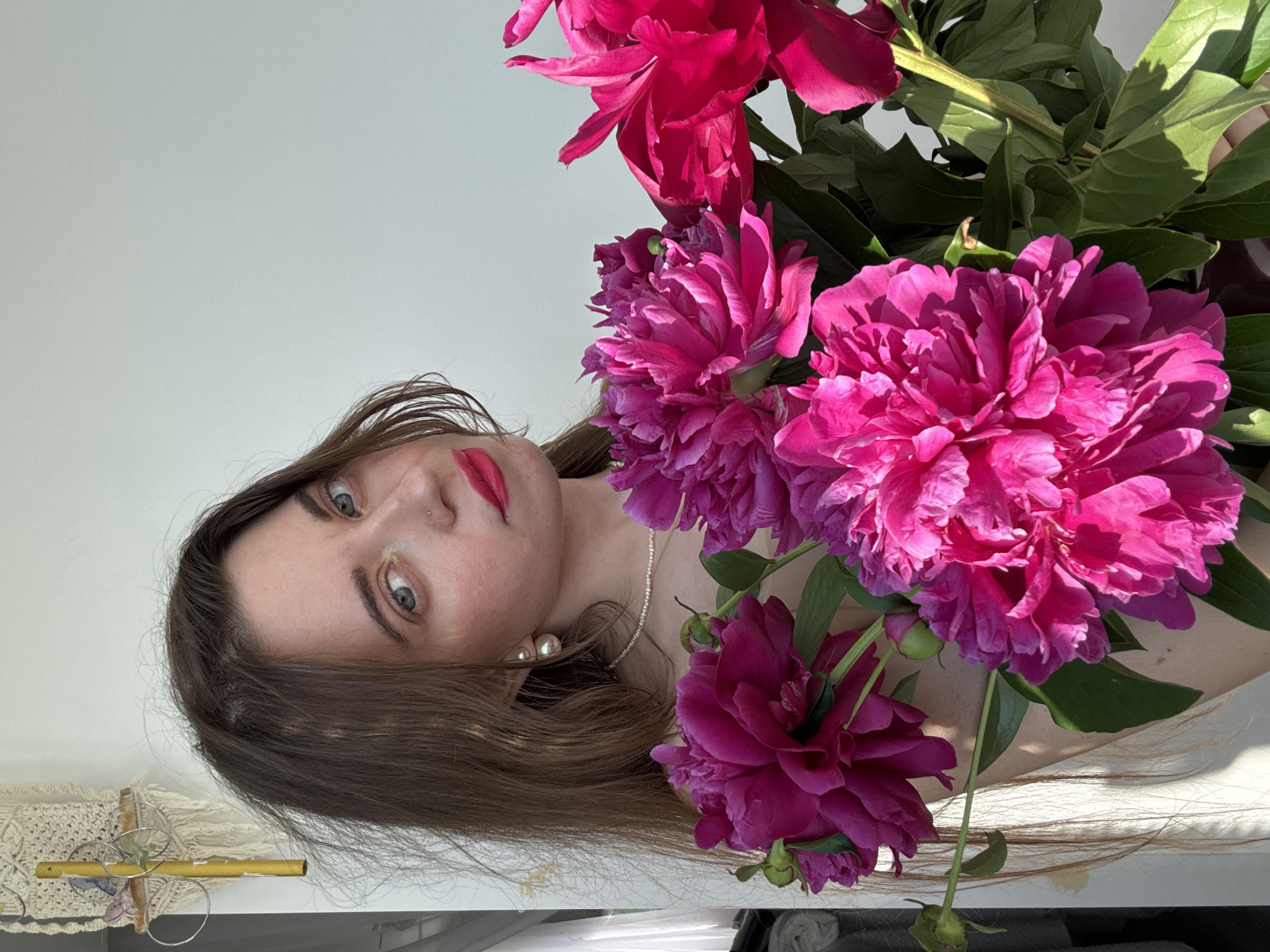 An over-the-shoulder, medium shot captures a woman in an indoor setting, holding a bouquet of bright pink peonies. The woman, with long brown hair, is positioned on the left side of the frame, her face turned towards the viewer. She wears a delicate pearl necklace and earrings. Her lips are adorned with vibrant pink lipstick, and her eyes, a striking blue, are looking directly at the camera. The peonies, the focal point of the bouquet, are in full bloom, their rich pink petals contrasting with the surrounding greenery. A macrame wall hanging with a gold cross, adorned with small crystals, is visible in the upper left background. The wall behind her is a plain, light color, reflecting the natural light streaming in from an unseen source. The lighting suggests it is daytime, with shadows cast by the flowers and the woman's hair. The setting appears to be domestic, possibly a home or studio, given the presence of the decorative wall hanging and the general ambiance.