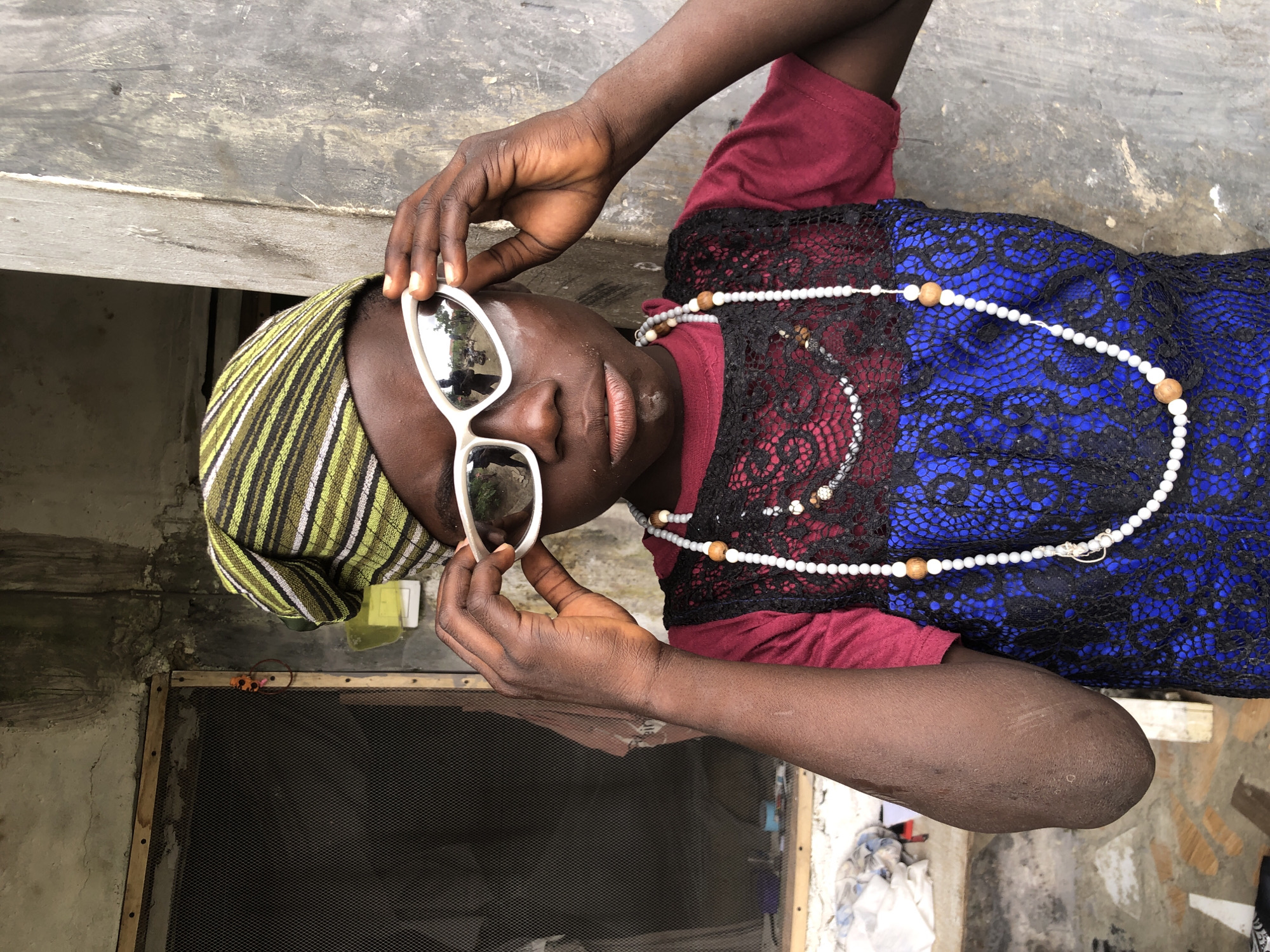 A person wearing a green and yellow striped headscarf and white-rimmed sunglasses is shown in a medium shot, standing in front of a textured gray wall. The person's hands are raised, holding the sunglasses in place. They are wearing a maroon shirt underneath a black lace garment with blue accents, and several beaded necklaces. White powder is visible on their face. A screened window is visible to the left of the frame, and the environment appears to be outdoors, possibly in a residential area of Ado, Nigeria, given the weathered construction materials and the overall aesthetic.