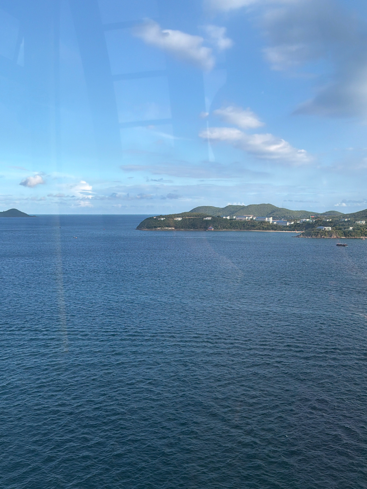 The image depicts a vast expanse of the ocean under a partly cloudy sky. In the distance, a lush green coastline with buildings is visible, suggesting a developed area. Two small islands are present in the sea. The water appears calm with a slight ripple. The presence of buildings on the coastline and the islands in the sea is consistent with the scenery around Nha Trang, Vietnam, a coastal city known for its beaches and islands.