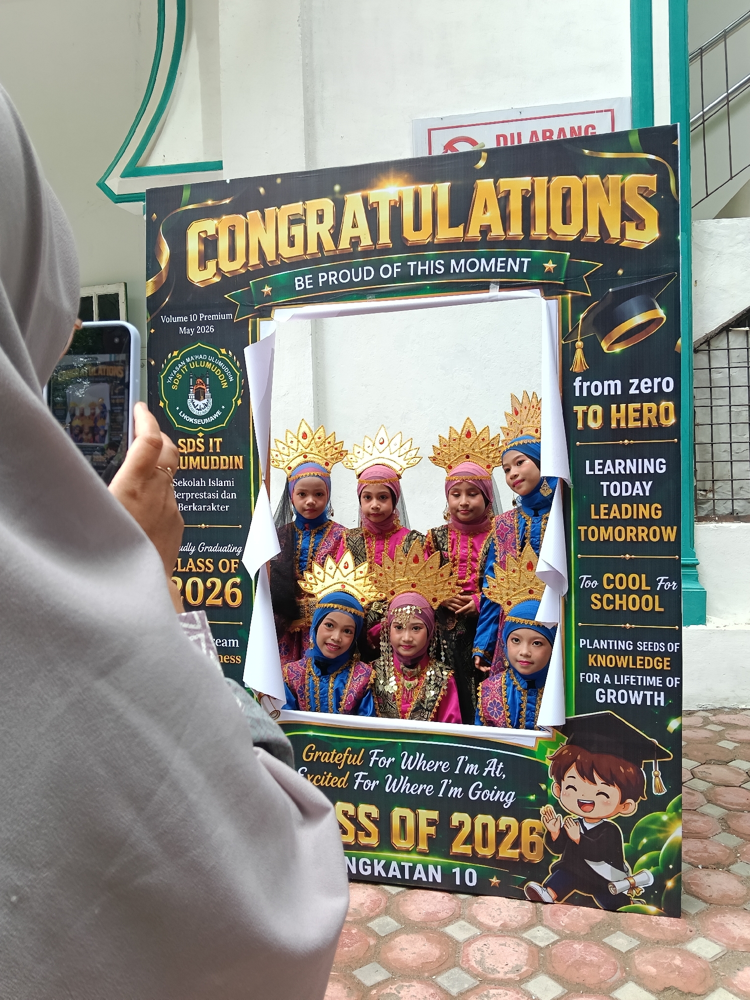 A person in a grey hijab is holding a phone and taking a picture of a graduation celebration photo booth. The photo booth displays "CONGRATULATIONS" and "BE PROUD OF THIS MOMENT." The backdrop is for SDS IT ULUMUDDIN, an Islamic school located in Lhokseumawe, Indonesia. The booth also has text such as "from zero TO HERO," "LEARNING TODAY LEADING TOMORROW," and "CLASS OF 2026." Through the cut-out window of the booth, seven young children, dressed in traditional attire and golden headdresses, are posing. The background indicates the photo is taken outdoors near a building.