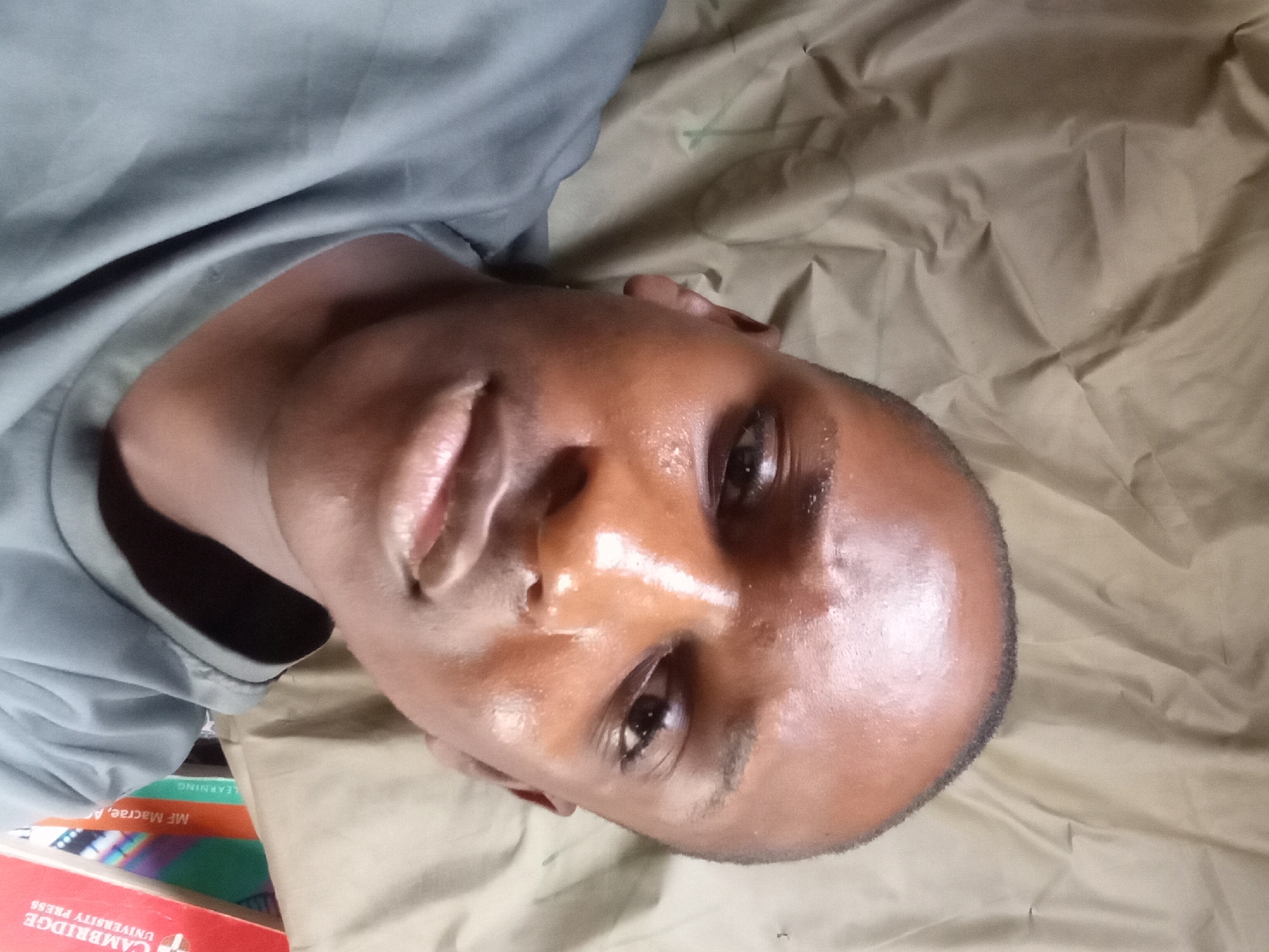 A close-up, eye-level, selfie-style photograph shows a young Black man with closely shaven hair, wearing a gray t-shirt. The man is positioned in the lower right quadrant of the frame, with his face turned towards the camera and his head tilted slightly. He appears to be in an indoor setting, as indicated by the presence of books stacked to his right. The background is a rumpled, tan-colored fabric, likely a curtain or bedsheet. The lighting in the image is somewhat harsh, creating highlights on the man's forehead and cheeks, suggesting sweat or oil on his skin. The scene appears to be a casual, personal environment. The image does not contain any specific visual cues that can be used to determine its location within Nkpor, Nigeria.