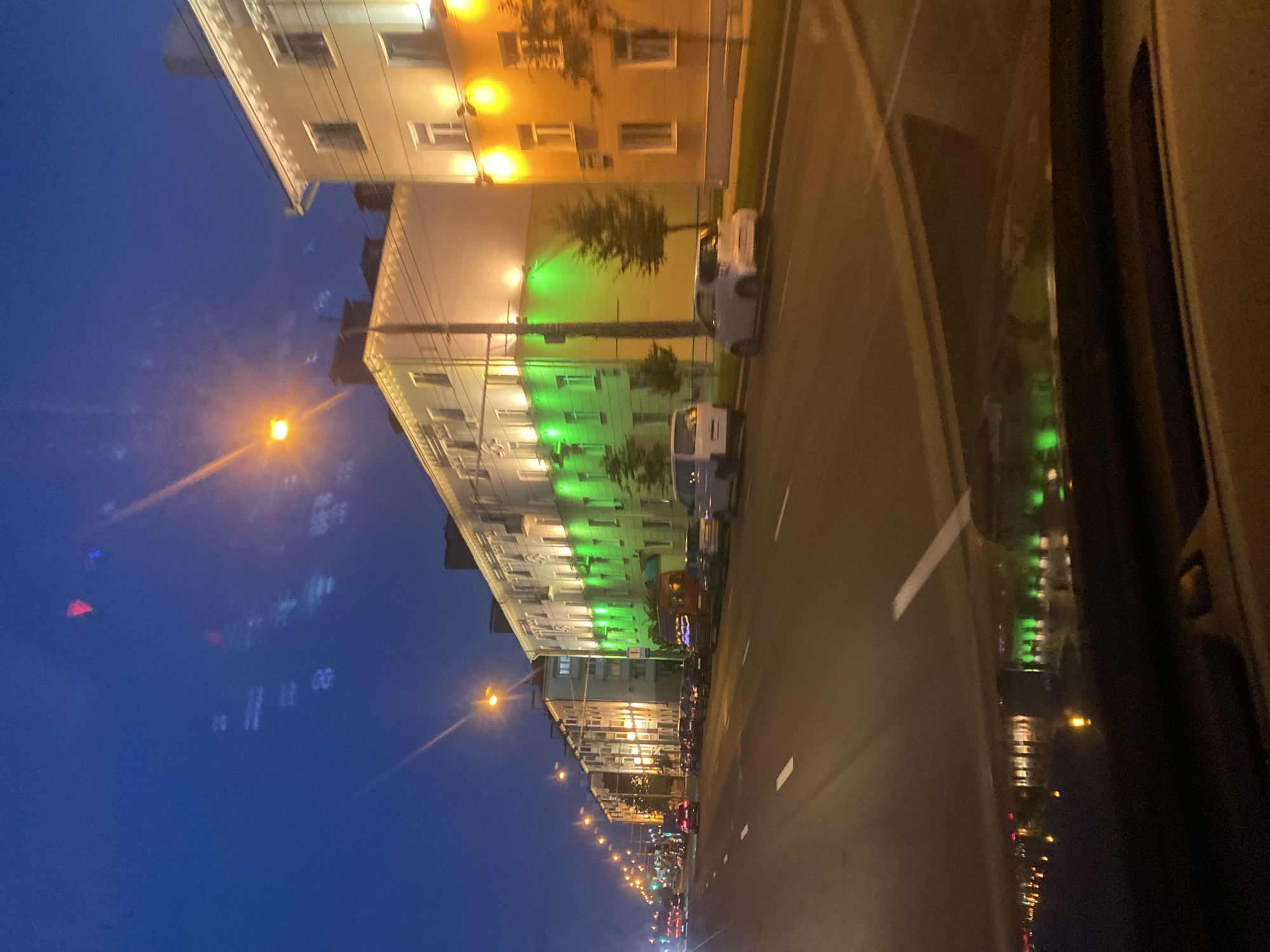 The image depicts a nighttime view from inside a vehicle driving along a wide city street in Minsk, Belarus. The street is lined with multi-story apartment buildings, many of which are illuminated with warm yellow and bright green lights. The sky is a deep blue, transitioning to a lighter shade near the horizon, with some wispy clouds visible. Streetlights line the road, casting a warm glow. Several cars are visible on the street, parked and in motion. A white van and a small white car are parked on the right side of the road. The reflection of the buildings and lights is visible on the wet surface of the road. The overall scene suggests a well-lit urban environment at dusk or night.