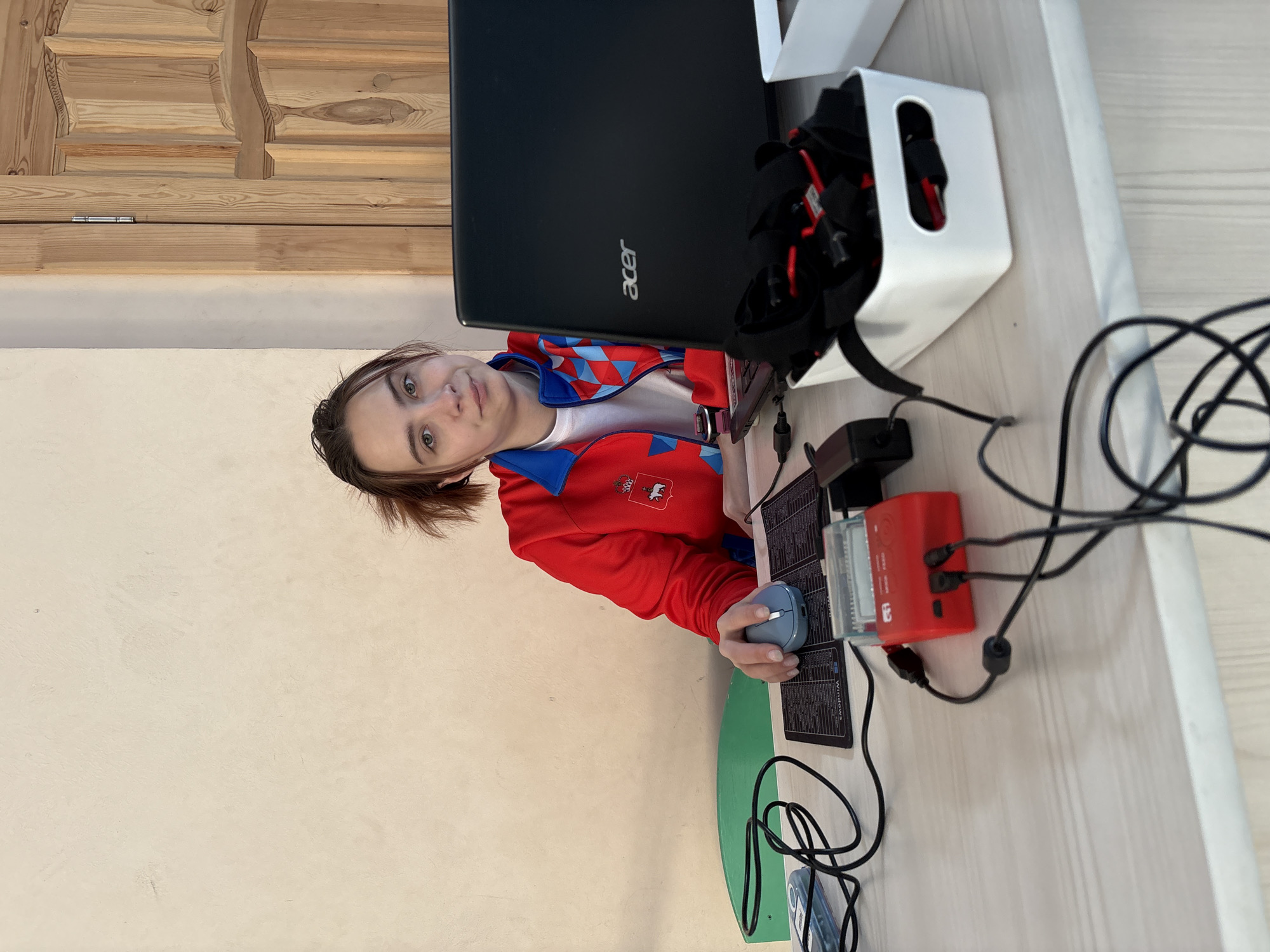 A person is seated at a desk with an Acer laptop, a white container holding red and black straps, a red electronic device, and a blue computer mouse. The person is wearing a red jacket with a white shirt underneath and has short brown hair. The desk is situated against a light beige wall and a wooden door. The image was taken indoors. I cannot confirm the location is in the city of Berezniki, Russia.