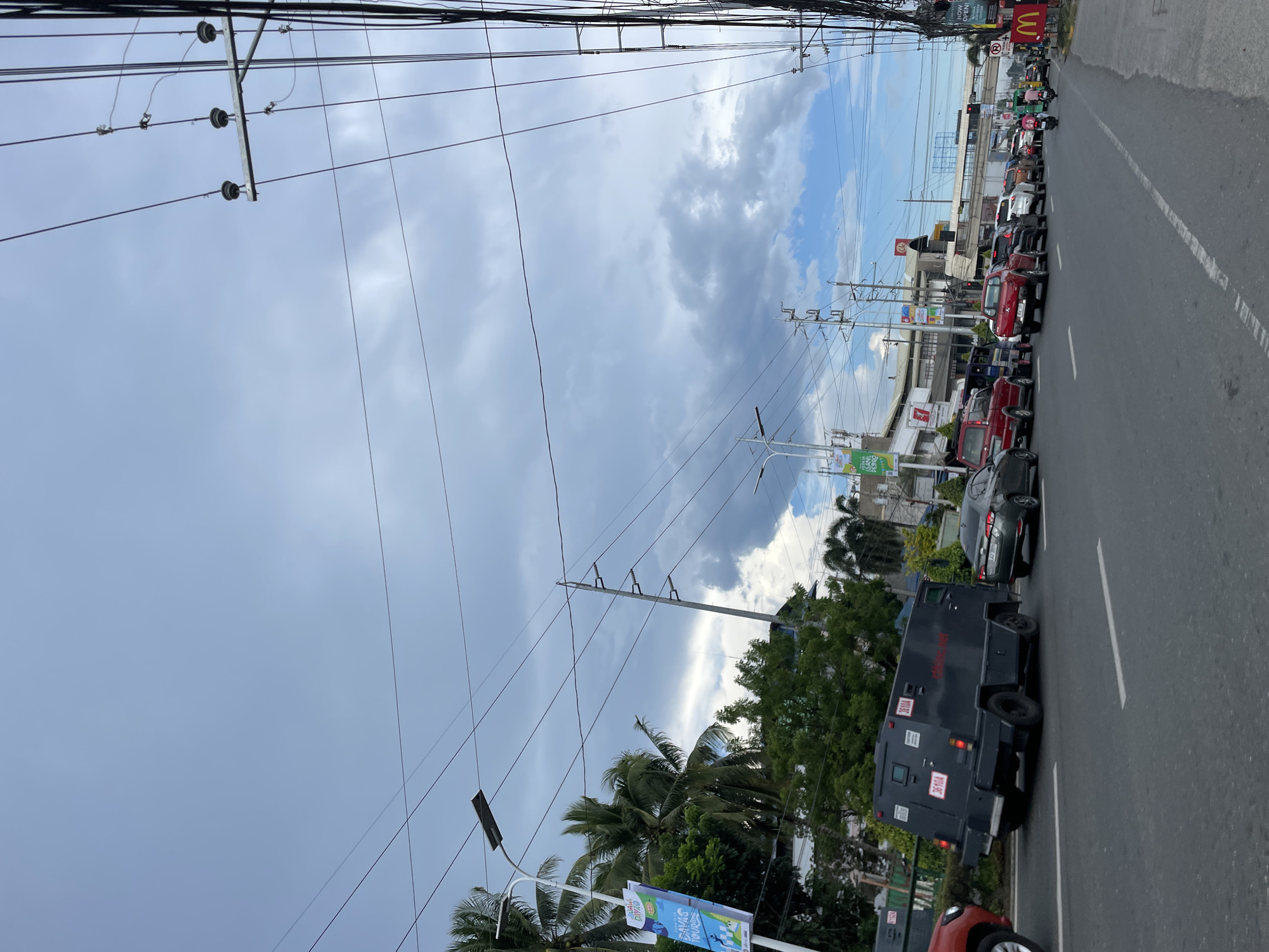 The image depicts a street scene in Davao City, Philippines, with a traffic jam on a cloudy day. In the foreground, a dark-colored armored vehicle with red markings is visible. Several other vehicles, including cars and a red pickup truck, are stopped in traffic. Palm trees and other greenery line the side of the road, and buildings with signage are visible in the background. Power lines and utility poles are prominent throughout the scene, indicating an urban or suburban environment. A banner with text in Filipino, likely advertising a local event or destination, hangs from a lamppost on the left. A McDonald's sign is visible on the right side of the road.