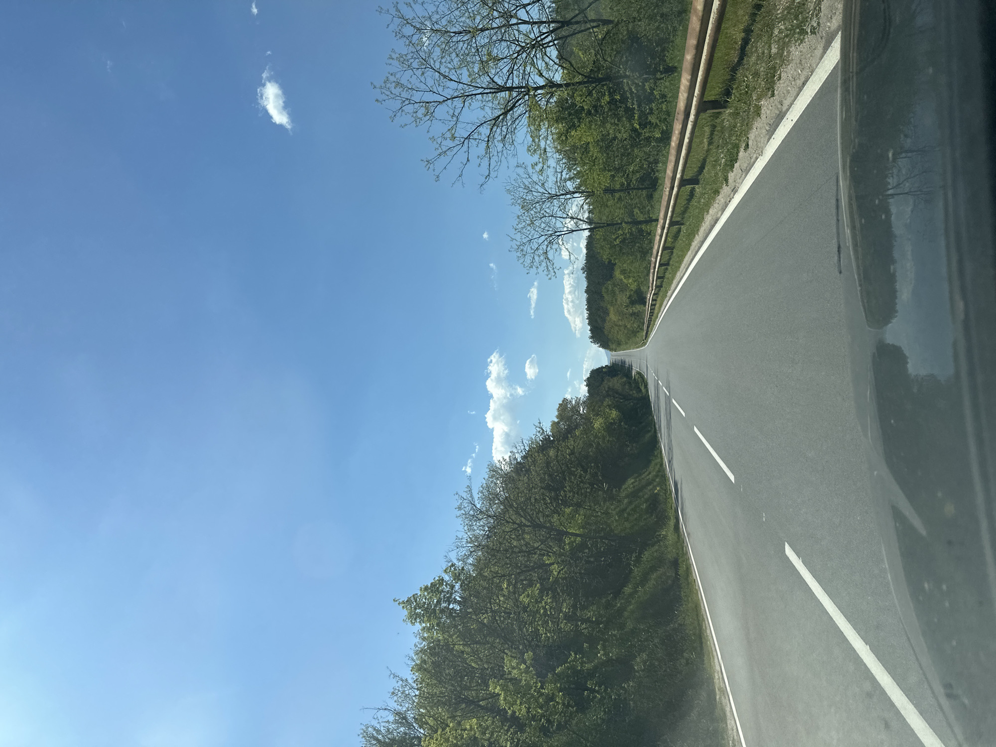The image shows a paved road running through a wooded area, under a clear blue sky with scattered clouds. A metal guardrail lines one side of the road. The location is unknown.