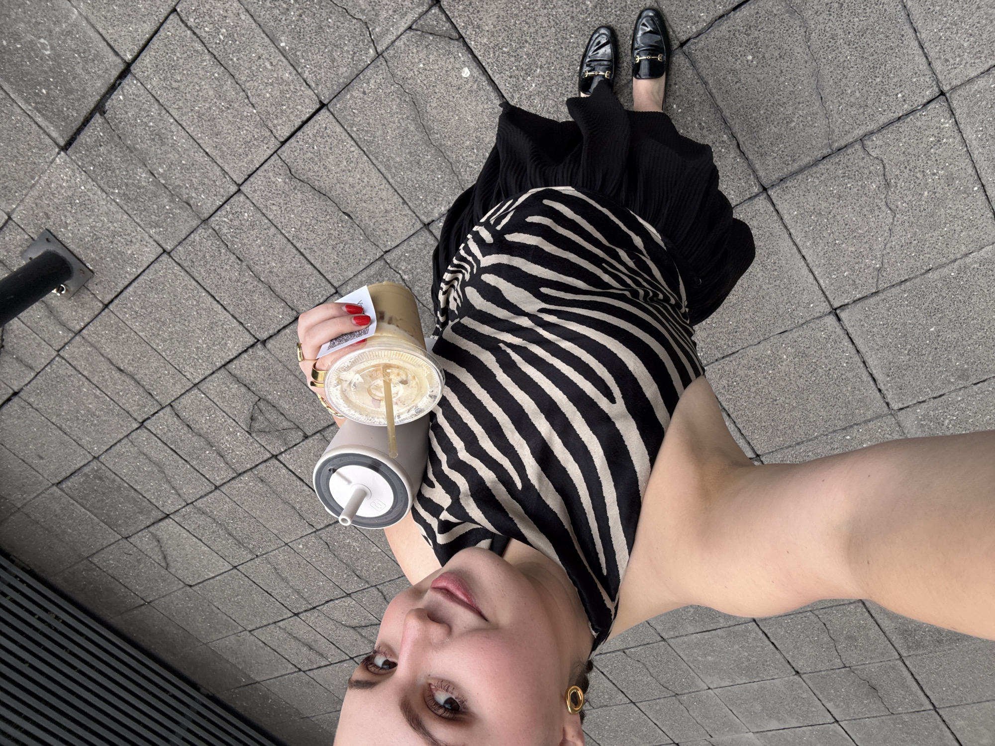 A woman is standing on a sidewalk made of grey rectangular paving stones. She is holding an iced coffee drink in her left hand and a reusable water bottle in the same hand. The woman is looking up toward the camera and taking a selfie. She has red fingernails, gold rings, and large gold earrings. She is wearing black loafers with gold horse-bit detailing, a long black pleated skirt, and a black and cream-colored zebra print top. A black metal pole can be seen in the background. The location is likely in Mexico City, Mexico.