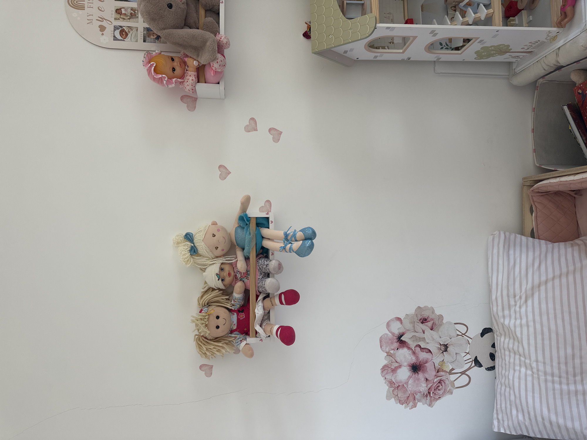 In a well-lit room, likely a child's bedroom in Petah Tikva, Israel, several items are arranged on and around a white wall. A narrow white shelf holds three cloth dolls. The doll on the left has blonde, frizzy hair and wears a red dress with white trim. The middle doll is smaller, with a light-colored hat and a pink shirt. The doll on the right has blonde hair tied with a blue bow and wears a blue tutu dress. The dolls' legs dangle over the edge of the shelf. Pink heart-shaped decals are scattered on the wall. To the right, a tiered white shelf displays a pink-bonneted doll and a large gray stuffed bunny. Above this shelf is a decorative arch with photos and the text "MY FIRST year". On the lower right, a multi-story dollhouse is visible, featuring green roof tiles and white walls with arched windows. Below the dollhouse is a light-colored hamper. In the lower left corner, a pillow with beige and white stripes lies on a striped bassinet. A floral wall decal with pink and white flowers is positioned above the pillow.