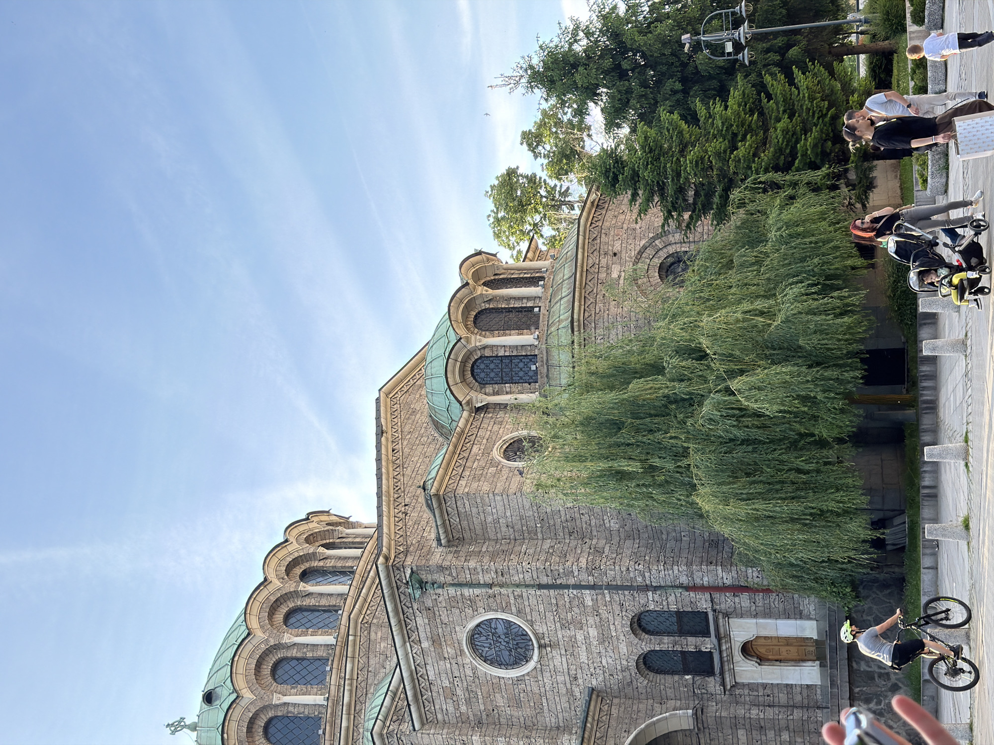 The image shows a section of the St. Alexander Nevsky Cathedral in Sofia, Bulgaria, viewed from a low angle. The cathedral's stone facade, arched windows, and copper-green dome are visible. A weeping willow tree partially obscures the building's facade.
Several people are present on the sidewalk. A cyclist wearing a helmet is riding a bike. Nearby, a woman with red hair pushes a stroller. A group of three people are standing and talking. Another person is walking away.
The sky is blue with some cloud streaks. The picture appears to be taken during the daytime with natural sunlight.