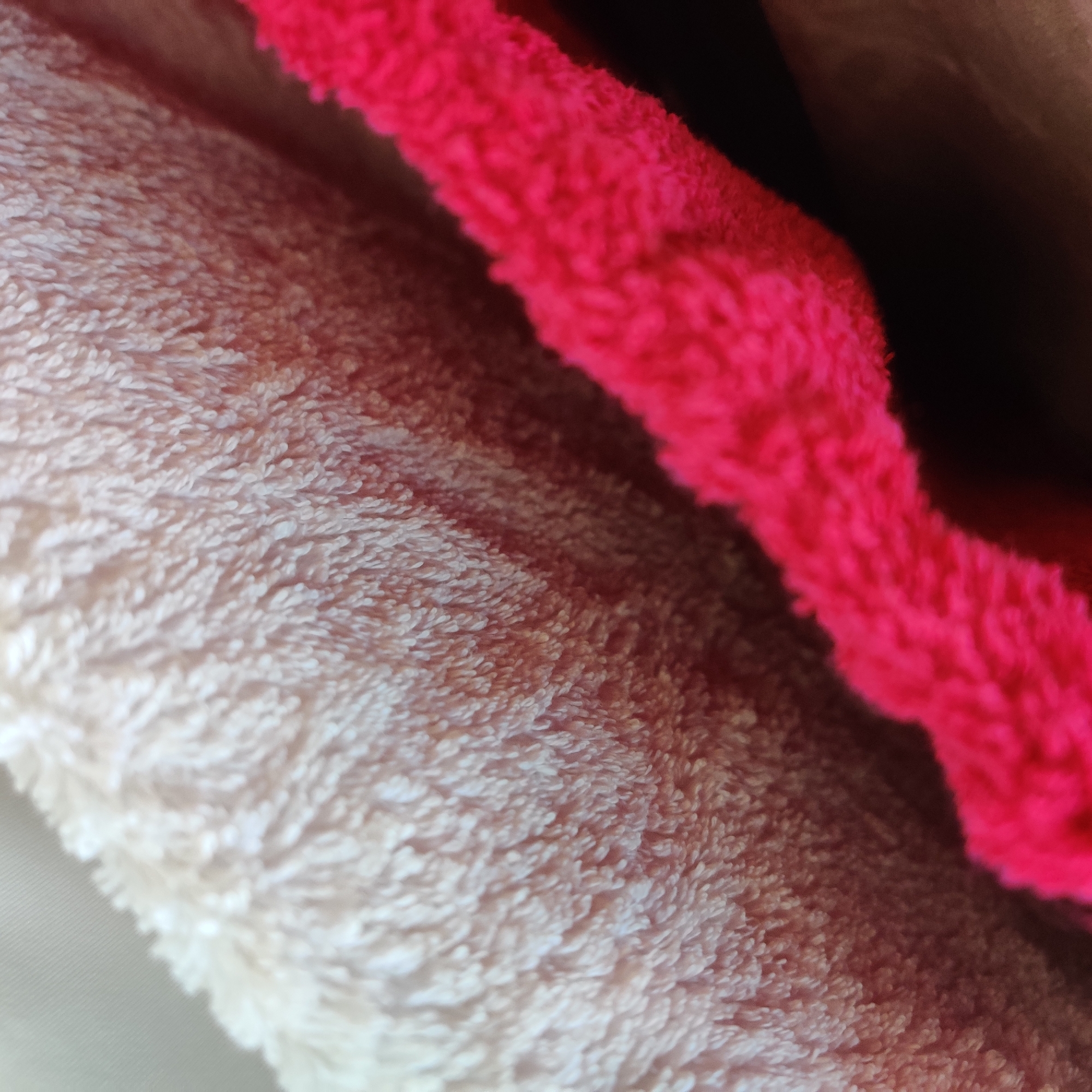 The image is a close-up, abstract shot of two plush fabrics. The fabric in the foreground is a vibrant hot pink, while the fabric in the background is a pale, creamy white. The textures of both fabrics are highly detailed, showing individual fibers and a soft, fuzzy appearance. The lighting is somewhat diffused, creating soft shadows that emphasize the three-dimensionality of the materials. The composition is diagonal, with the pink fabric running from the lower right to the upper center of the frame, creating a dynamic visual flow. The background is out of focus, drawing attention to the textures and colors of the fabrics. The image does not contain any elements that would allow for identification of a specific location in Yekaterinburg, Russia.