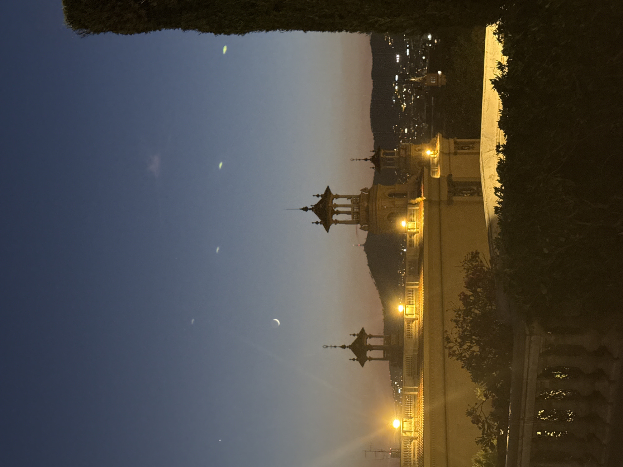The image captures a twilight view of the National Palace (Palau Nacional) in Barcelona, Spain, as seen from the Montjuïc hill. The sky transitions from a deep blue at the top to hues of orange and pink near the horizon, indicating the end of daylight. The Palace, a prominent landmark, is illuminated with warm yellow lights along its facade and on decorative lamps lining a balcony. Several architectural elements of the Palace are visible, including ornate towers and columns. In the distance, the city of Barcelona glows with numerous lights, set against a dark mountain range. The crescent moon is visible in the sky. A tall, dark evergreen tree stands on the right side of the frame, partially obscuring the view of the city lights. In the foreground, dark foliage and a stone balustrade create a sense of depth and frame the scene. The overall atmosphere is serene and picturesque, characteristic of a city view at dusk.