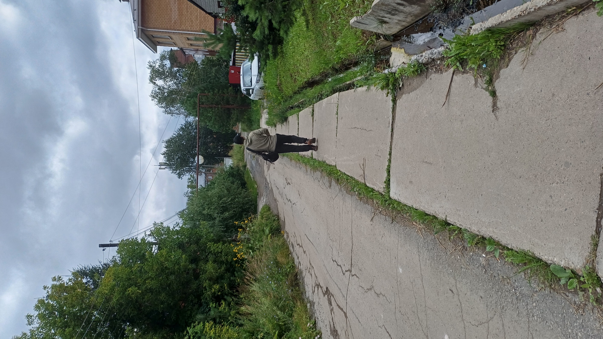An individual wearing a light brown hooded jacket, dark pants, a black baseball cap, and brown shoes walks away from the camera on a concrete sidewalk. The person carries a black bag over their left shoulder. The sidewalk is bordered on the left by a cracked asphalt road and on the right by a drainage ditch with overgrown grass. Beyond the ditch, a white car is parked next to a brick building with a pitched roof. Trees and vegetation are abundant on either side of the road, with power lines and utility poles visible in the background under a cloudy sky. The scene appears to be in a residential or semi-rural area of Izhevsk, Russia.
