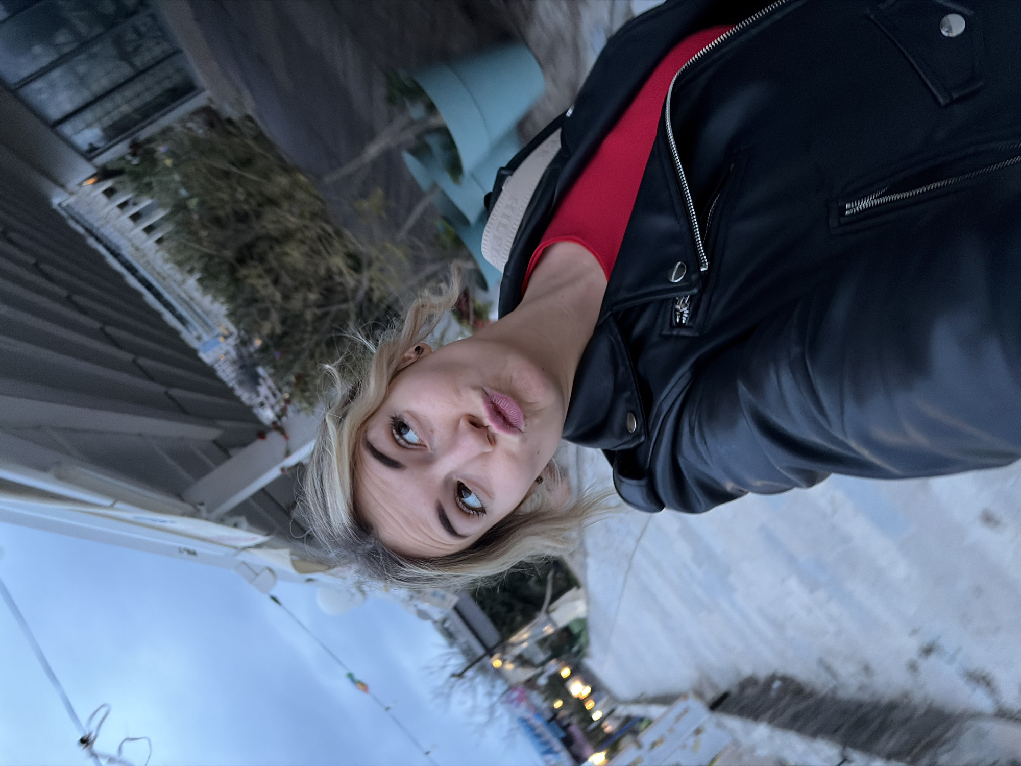 A fair-skinned woman with short blonde hair poses in front of what appears to be a holiday market in Budva, Montenegro. She is wearing a red shirt and a black leather jacket. The background shows a sky with a light blue hue, suggesting twilight, and blurred lights, likely from the market. The market stall roofs are visible. There are also blue pots in the background. The image is taken at an odd angle.