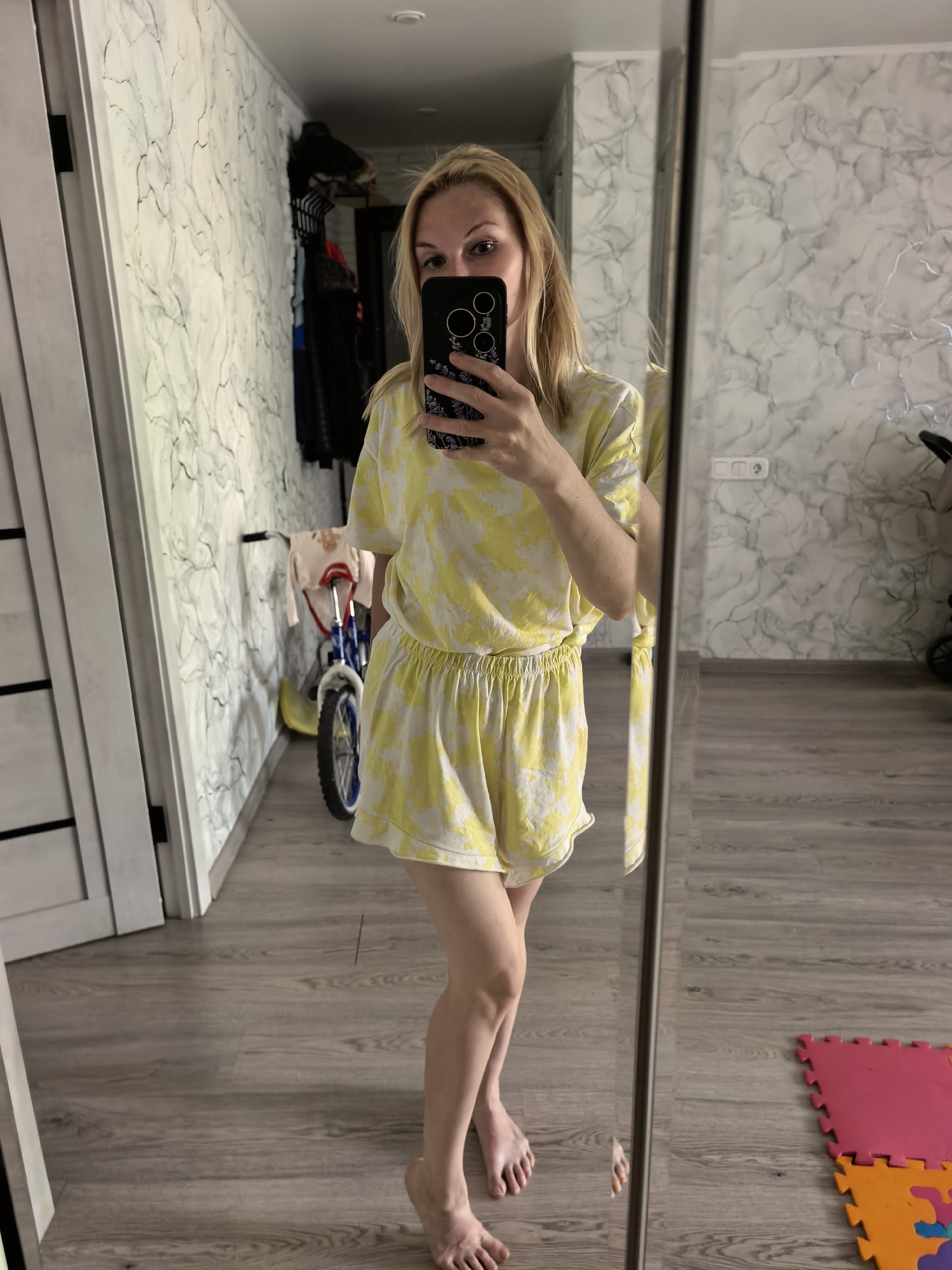 A fair-skinned woman with blonde hair is taking a mirror selfie in an indoor setting, likely a home. She is wearing a matching yellow and white tie-dye shorts and shirt set. She holds a black smartphone in front of her face, partially obscuring it.
The background includes a hallway with light gray wood-look flooring. The wall on the left has a marble-like wallpaper. There is a doorway on the left. Behind the woman, a child's bicycle is partially visible. Further back, there are clothes hanging in what appears to be a closet or storage area.
To the right of the woman, the mirror reflects a portion of the room, including more of the marble-like wallpaper and what appears to be a stroller. The floor in this area has interlocking foam mats, colored in pink, orange, and purple.