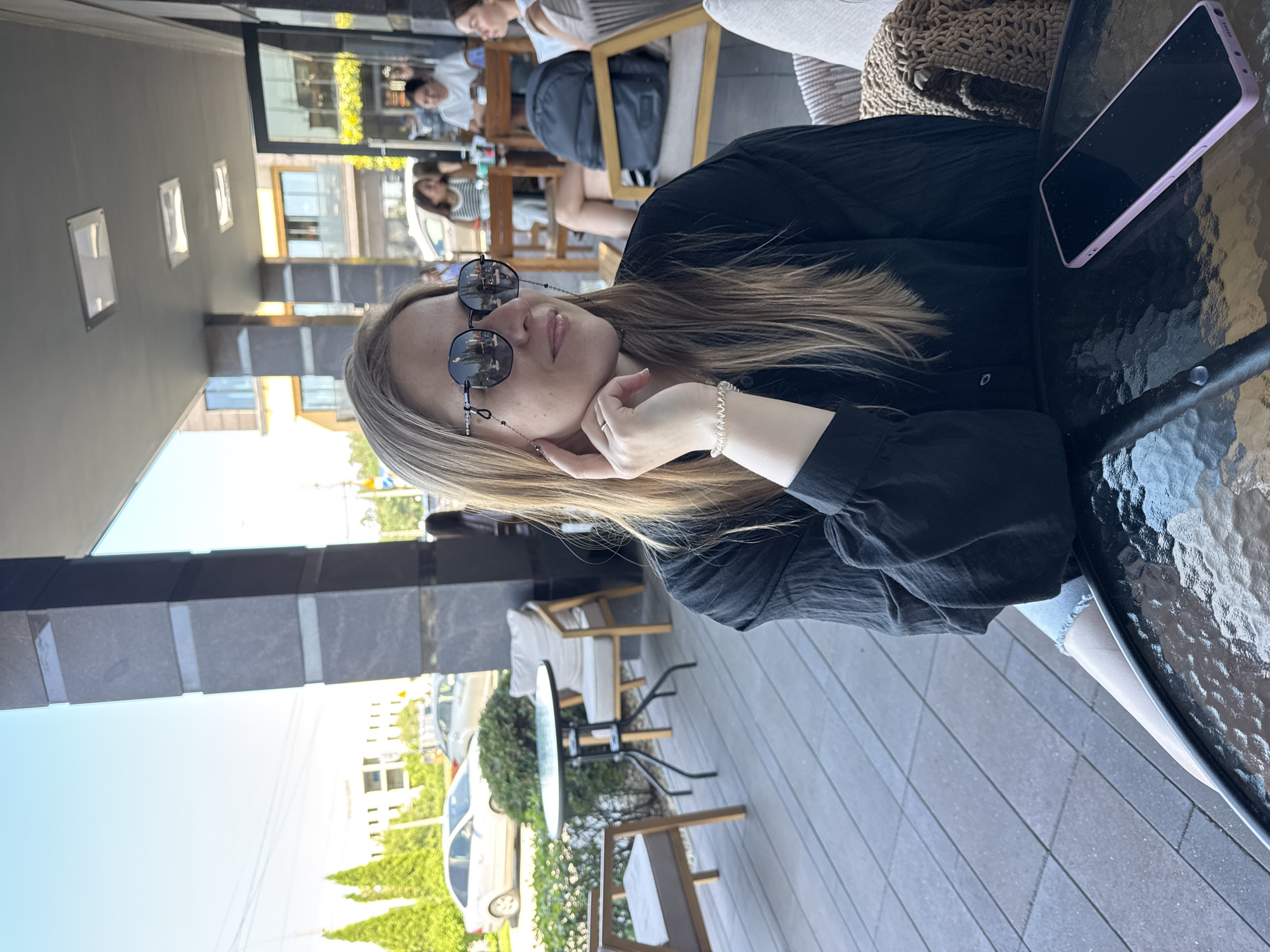 In an outdoor seating area, a woman with blonde hair and sunglasses sits at a black glass-top table. She is wearing a black long-sleeved shirt and denim shorts. Her left hand rests on her chin, and a white bracelet is visible on her wrist. A smartphone in a pink case is on the table in front of her. In the background, other patrons are seated at tables and chairs, and trees and buildings are visible. The flooring is a grey paved patio.