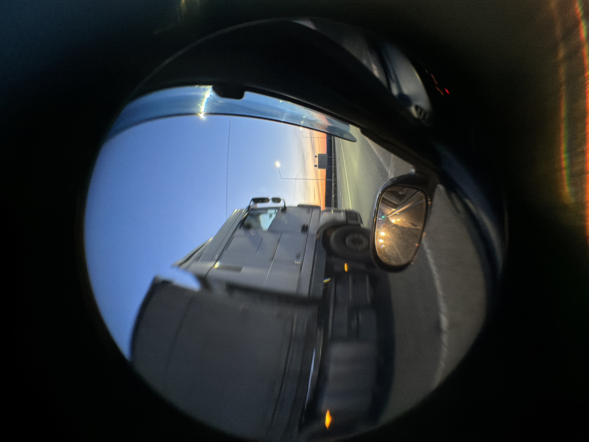 A fisheye-lens perspective from inside a vehicle shows a large white truck passing on a highway. The sky is visible with the last vestiges of a sunset, and streetlights are illuminating the road. A side mirror is also visible. The location is not discernible from the image.