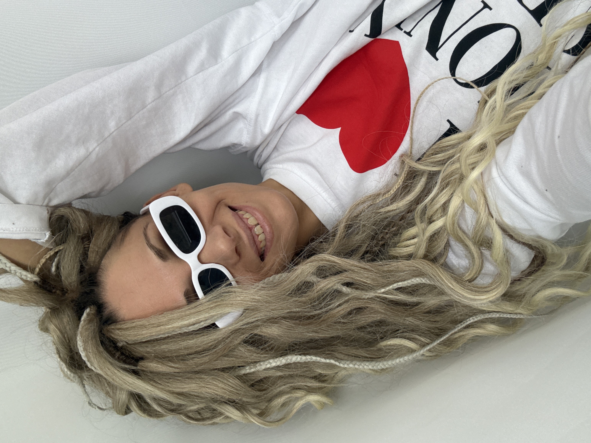 The image depicts a woman lying down, wearing a white long-sleeved shirt with a red heart and the word "NO" printed on it. She is also wearing white framed sunglasses. Her hair is long, wavy, and a mix of blonde and brown, with some sections braided with white thread. The background appears to be a plain white surface. There's no indication of the location being in Rusanovo, Russia, or any other specific location.
