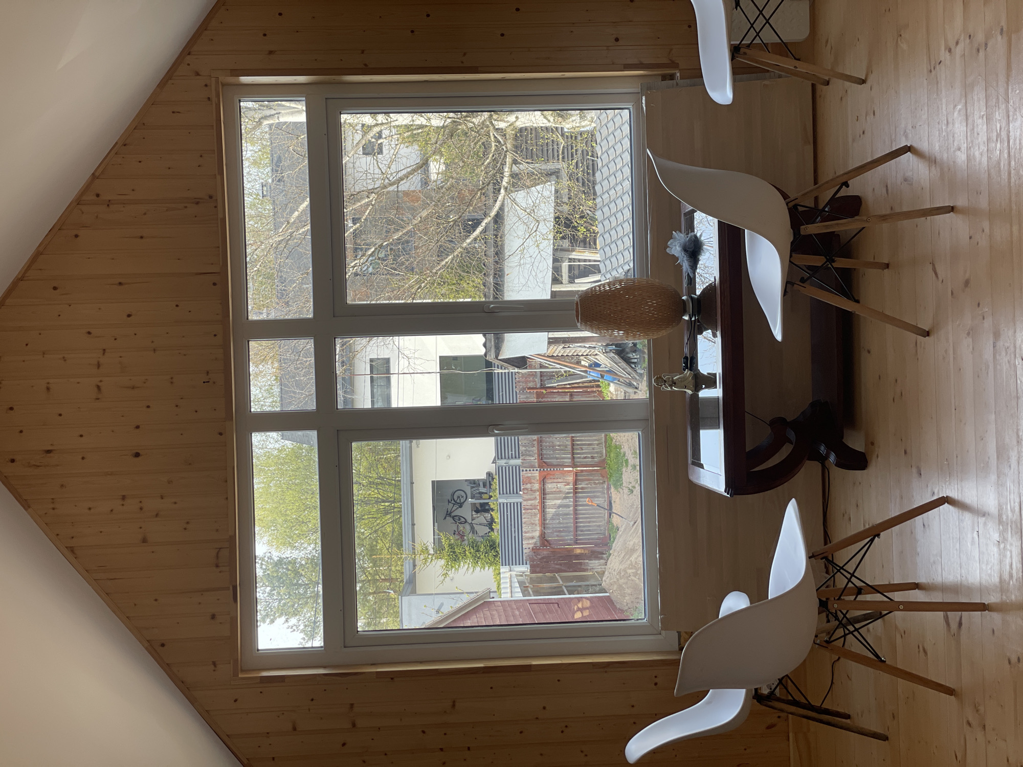 The interior of a room, likely an attic, is visible. The room has wood-paneled walls and a wooden floor. A large, multi-pane window dominates the frame, offering a view of an outdoor residential area. The outdoor scene includes buildings, trees with early spring foliage, fences, and what appears to be a bicycle. There are a table and three white chairs with wooden legs in the room. The table has a decorative lamp and a feather duster on it. The location is likely Sestroretsk, Russia, based on the architecture and vegetation. There are no people visible.