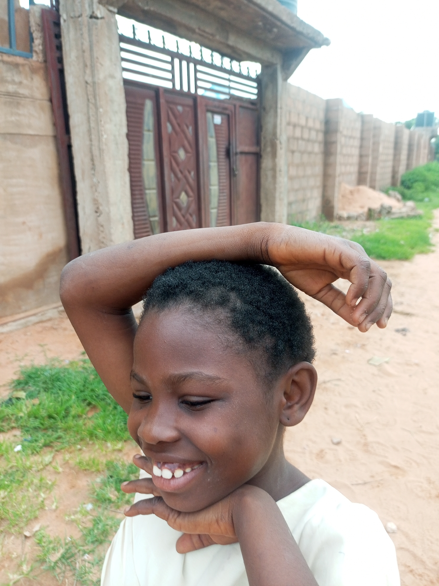 The image depicts a young Black person, likely a child, with dark skin and short, dark, curly hair. The person is smiling broadly, revealing their teeth, and has their right arm raised, with their hand resting on their head and their fingers loosely clasped. Their left hand is positioned near their chin. They are wearing a light-colored t-shirt.
The background suggests an outdoor, possibly semi-urban or rural, environment in Tempure, Nigeria. To the left, there is a light-colored concrete structure with a rough texture, and behind it, a dark metal gate with decorative elements. A brick wall, constructed from light brown bricks, extends to the right and recedes into the distance. The ground is covered with sandy soil and patches of green grass. In the distance, more brick walls and some vegetation are visible, along with what appears to be a water tank on a rooftop. The sky is overcast and bright. The overall scene appears to be a snapshot taken in a residential or community area.