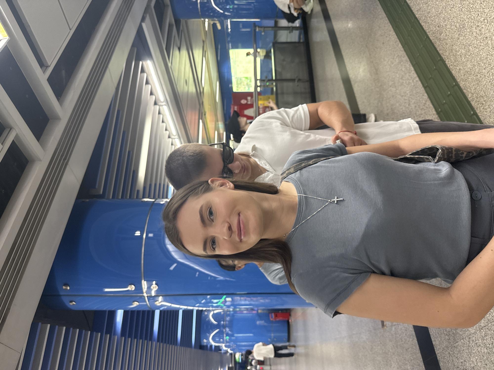 In Haizhu District, China, a medium shot captures a young Caucasian woman, center-frame, looking directly at the camera with a slight smile. She has long, dark brown hair, blue eyes, and is wearing a grey t-shirt and a silver cross necklace. To her right, a Caucasian man with short brown hair and sunglasses, wearing a white polo shirt, is partially visible, looking down. Behind them, other individuals are blurred and out of focus, one wearing a light grey shirt. The background features a modern subway station with prominent blue cylindrical columns, a suspended ceiling with fluorescent lighting, and a patterned floor with a tactile paving strip. A red trash can is visible to the left. The overall scene suggests a public transportation environment.