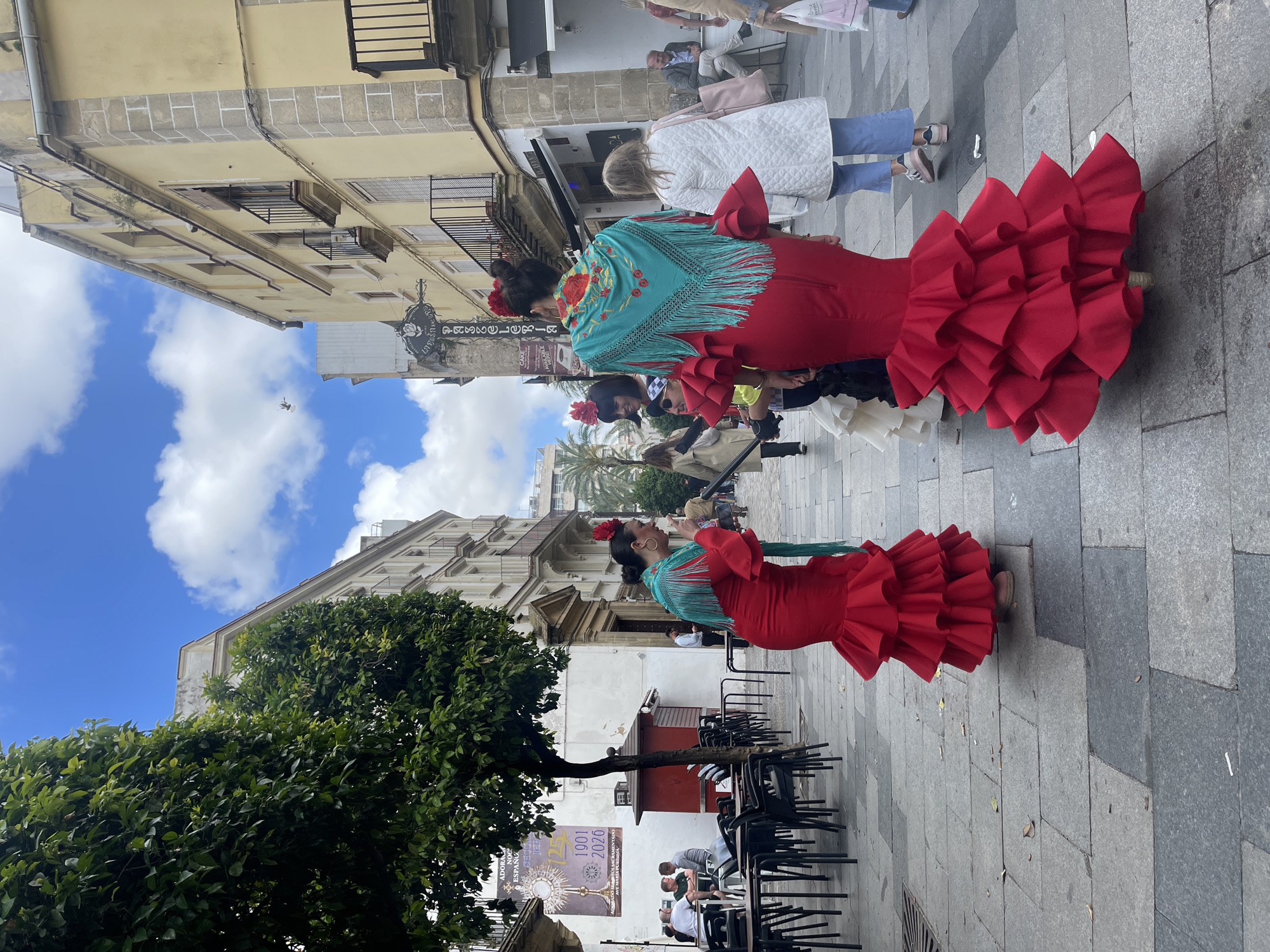 The image captures a street scene in Jerez De La Frontera, Spain. Two women are prominently featured, wearing vibrant red flamenco dresses with ruffled skirts and turquoise shawls adorned with floral embroidery. Both have their hair up with a red flower. They are standing on a paved street with a backdrop of buildings with a bright blue sky and white clouds above. A large green tree is to the left of the lower figure. The street appears to be a pedestrian zone with people walking in the background, and there are tables and chairs of an outdoor cafe in the foreground.