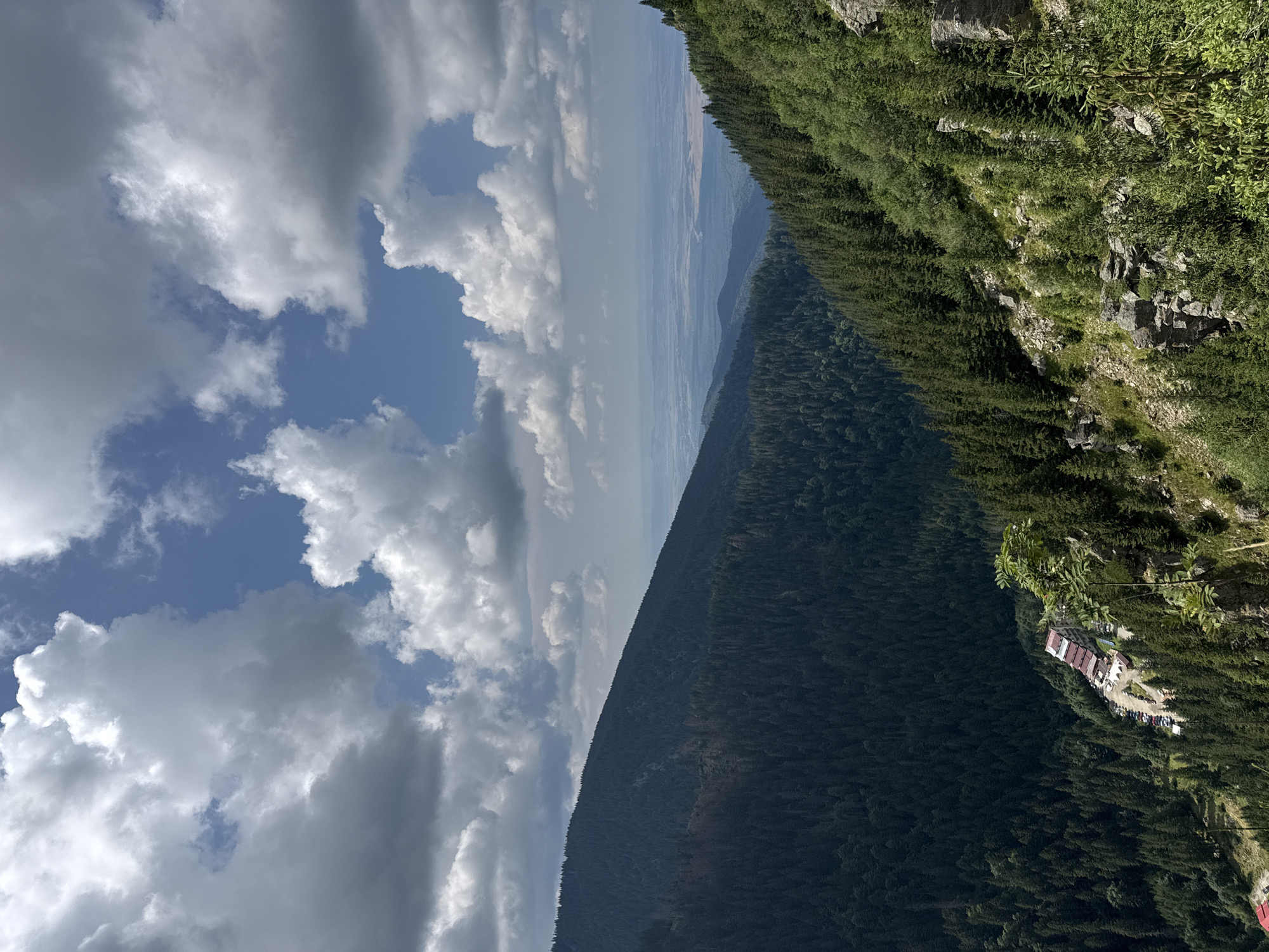 The image shows a mountainous landscape with a dense forest of evergreen trees covering the slopes. A building complex with red roofs and a parking lot is visible nestled in a valley. The sky is partly cloudy with dramatic white and gray clouds against a blue sky. In the distance, a vast, hazy plain stretches to the horizon. This is likely the Transfăgărășan mountain road, a popular tourist route in Romania, near Cârțișoara, with the building potentially being a guesthouse or restaurant. The scene is dominated by natural elements, with human presence indicated only by the man-made structures.