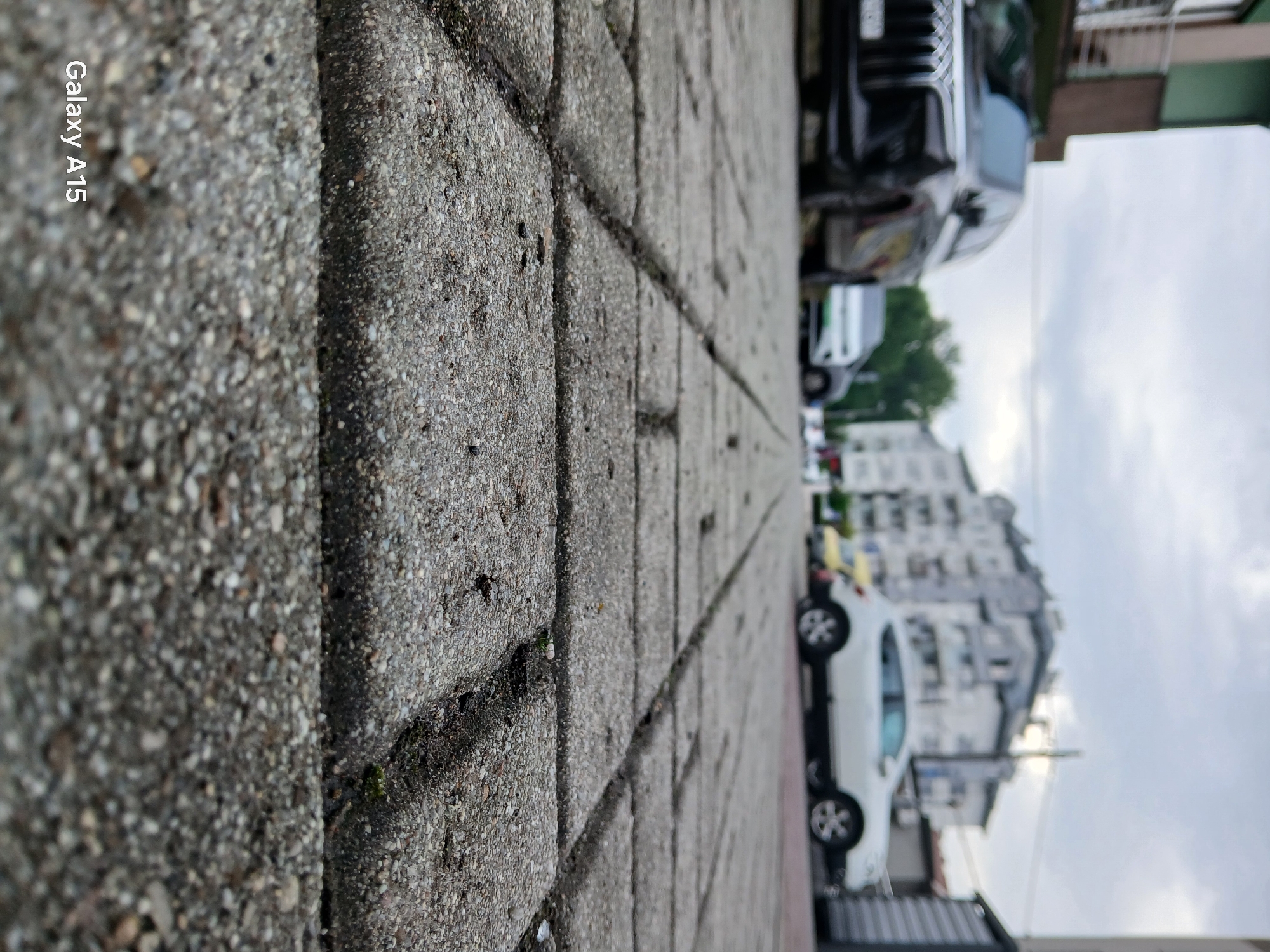 The image is a low-angle, Dutch tilt shot of a sidewalk paved with gray, rough-textured concrete blocks. The foreground is dominated by the detailed texture of the sidewalk, with small pebbles and cracks visible. To the right of the sidewalk, a street is visible with several cars parked and one white car moving, its wheels turned. The cars are dark and modern, suggesting an urban or suburban setting. In the background, there are buildings with multiple stories, appearing to be apartments or businesses. The sky is overcast and a pale gray, indicating a cloudy day. The time of day is difficult to determine precisely due to the lighting conditions, but it appears to be daytime. The overall mood is neutral, with no strong emotional cues. The text "Galaxy A15" is visible in the upper left corner of the image, indicating the device used to capture the photograph.