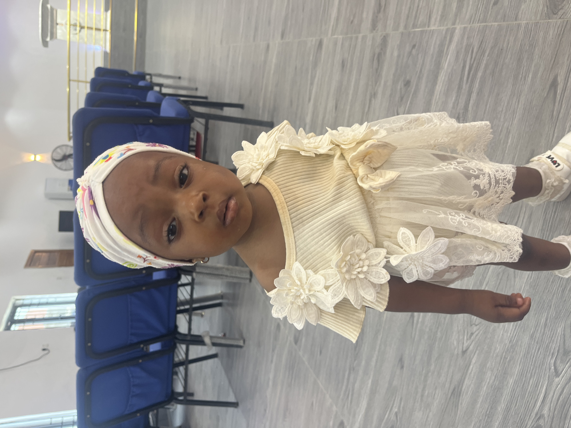 A young Black girl, appearing to be under five years old, stands in what appears to be a church or assembly hall in Ado, Nigeria. She is wearing a cream-colored dress adorned with lace flowers and a matching headband. Her expression is neutral. Behind her, rows of blue chairs are visible, and in the background, a podium and a wall with decorative lighting can be seen. The floor is tiled with a wood-like pattern. The lighting suggests an indoor setting, possibly during daytime.