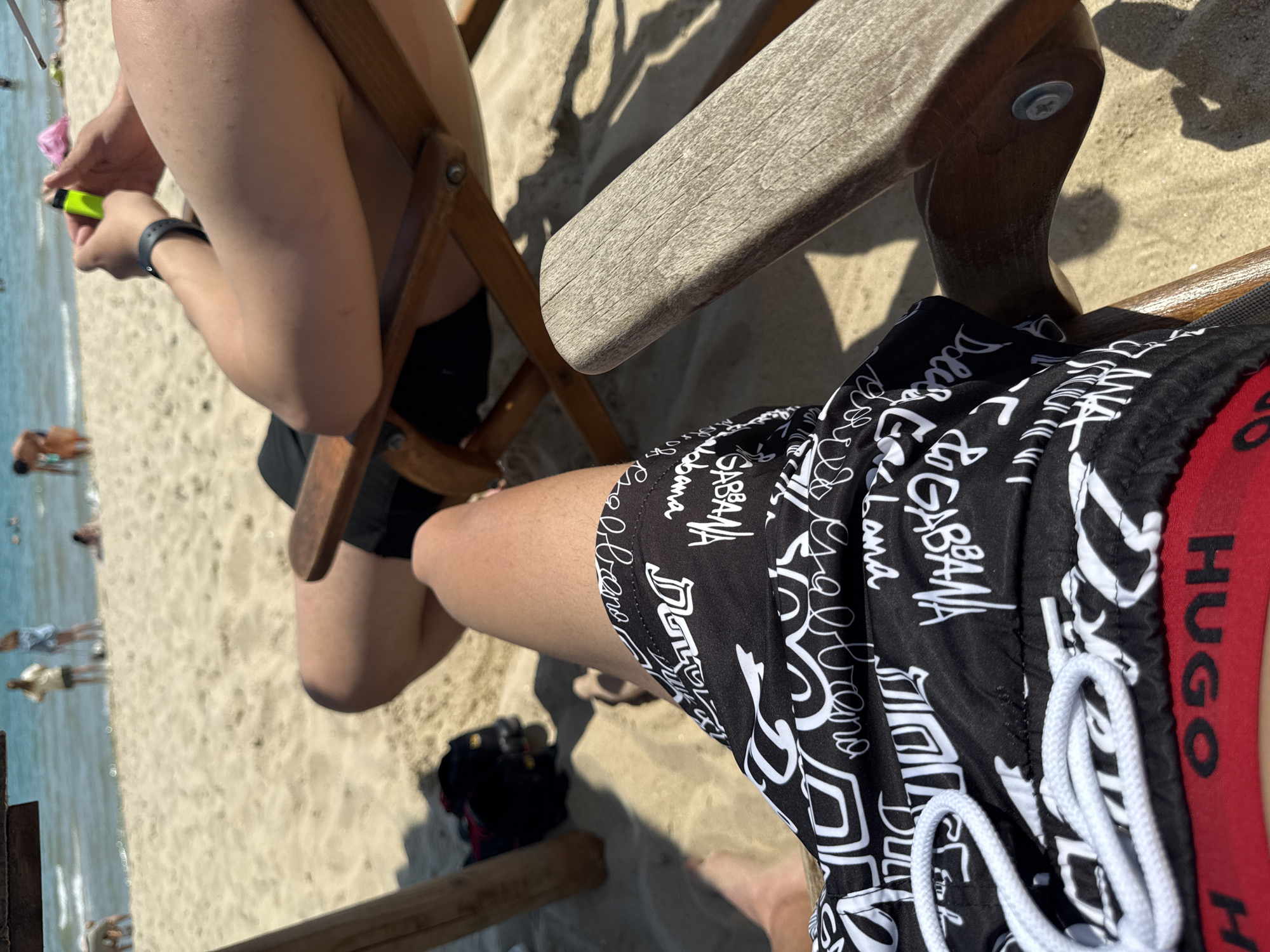 The image depicts a first-person perspective of a sunny beach scene in Varna, Bulgaria, viewed from a seated position in a wooden beach chair. In the immediate foreground, the photographer's tanned legs are visible, wearing black swim shorts adorned with white stylized text, including identifiable "Dolce & Gabbana" branding, with a white drawstring at the waist. Beneath the shorts, a red waistband featuring "HUGO" in black lettering is partially visible. The weathered wooden armrest of the chair extends into the frame on the right, while a wooden support column is present on the left.
In the midground, another individual is seated in an adjacent wooden beach chair, facing away from the photographer. This person, with tanned skin, wears black swim shorts and is holding a bright neon green object, likely a lighter, in their hands. A black wristband is visible on their left wrist. A black bag, revealing a red interior, rests on the light-colored sand to the left of the second person. Strong shadows from the chairs and individuals indicate direct overhead sunlight.
The background shows a wide expanse of light, fine sand extending towards the calm, blue-green sea. Multiple people are present on the beach and in the water. Figures include two women standing near the shoreline, another person bent over near the water's edge, and several individuals wading or swimming further out in the sea. The scene conveys a leisurely atmosphere on a clear, bright day at a coastal location in Varna, Bulgaria.