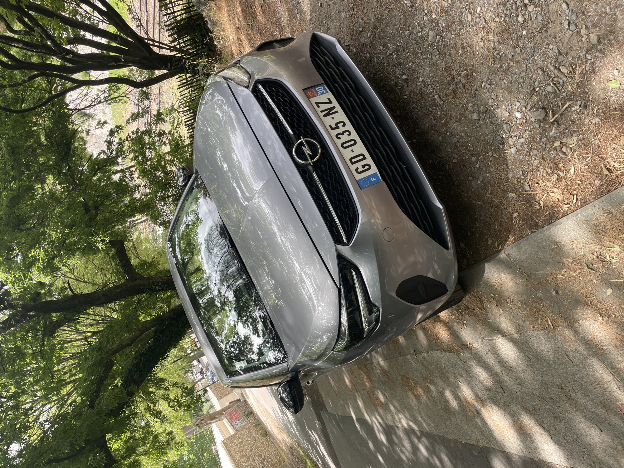 A silver Opel car is parked on the side of a gravel road with dry leaves and pine needles scattered on the ground. The car is positioned at an angle, with its front facing towards the right side of the frame. The sun is casting dappled shadows from overhead trees onto the car and the road, suggesting it is daytime. The scene is framed by lush green trees with dense foliage on the left and in the background. A black metal fence runs along the top left of the image, bordering some railway tracks that are partially visible. The car's license plate is visible and reads "GD-035-NZ". The overall impression is a quiet, outdoor scene, possibly in a rural or semi-rural location, with the car parked amidst natural surroundings.