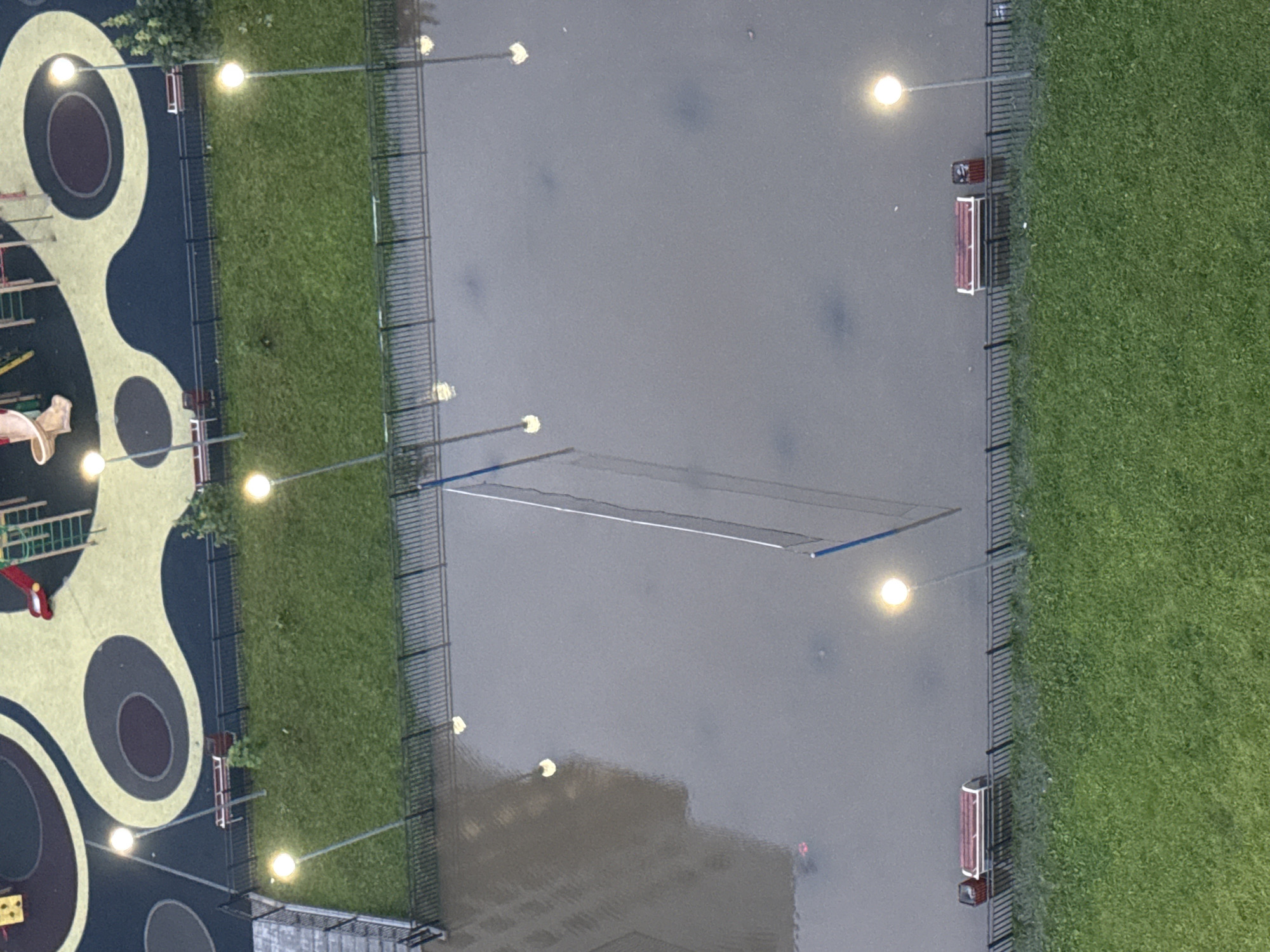 This is a high-angle, overcast, outdoor photograph of a flooded volleyball court in the city of Sinyavino, Russia. The court is filled with murky water, reflecting the light from street lamps. A volleyball net is set up in the center of the court. There are benches and trash cans along the perimeter fence, some of which are partially submerged. To the left of the court is a building, and to the right is a grassy area with trees. In the upper portion of the image is a playground with colorful rubber surfacing. The scene appears to be a residential courtyard after heavy rainfall.