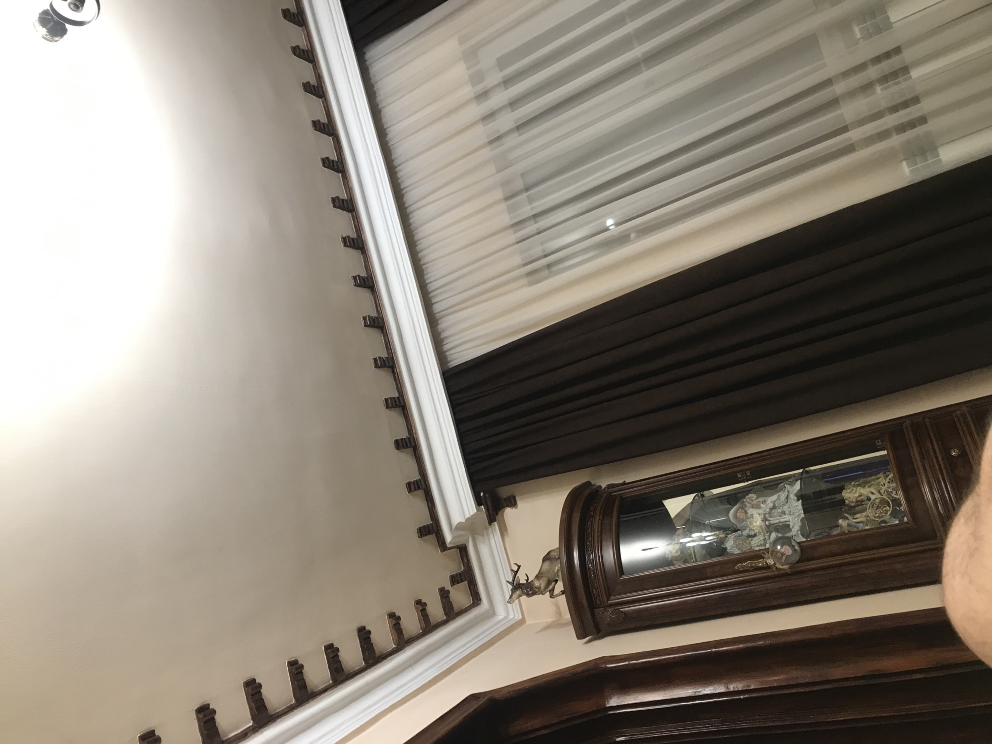 The image presents an interior shot, likely taken at an upward angle. The wall is painted in a pale beige, and a decorative molding adorns the corner where the wall meets the ceiling. The molding features a repetitive pattern of what appears to be small, evenly spaced brackets.
A window is visible at the top of the frame, covered with a layered window treatment. The inner layer consists of sheer, light-colored blinds, while the outer layer is a darker, brown curtain.
On the wall, to the right side of the frame, hangs a wooden-framed decorative item. Attached to it is a statuette of a stag.
The image does not provide enough information to determine the location as Yerevan, Armenia, or any other specific place. There are no discernible landmarks, signs, or other identifying characteristics. There are no people visible.