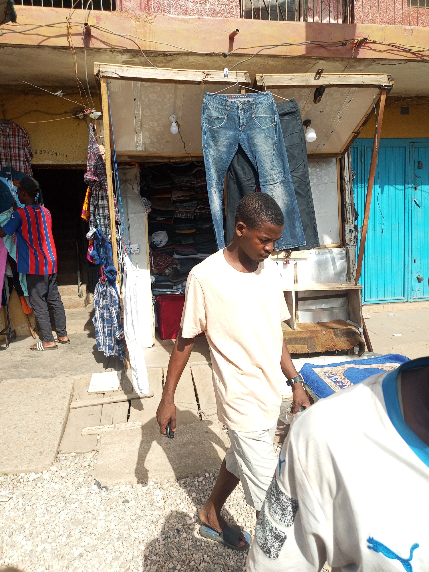 The image depicts a street scene in Maiburiji, Nigeria. In the foreground, a young man wearing a light pink T-shirt and white shorts walks to the right, holding a small, dark object in his left hand. He wears sandals and a wristwatch. Another person, wearing a white and blue shirt, is partially visible in the lower right corner.
Behind the first man, a small outdoor clothing stall displays items like jeans and shirts. A second man, wearing a red and blue striped shirt and dark pants, stands near the stall, examining the merchandise. The stall is situated in front of a building with a faded yellow facade. A pair of light blue doors are visible to the right of the stall. The ground is a mix of concrete slabs and gravel.
