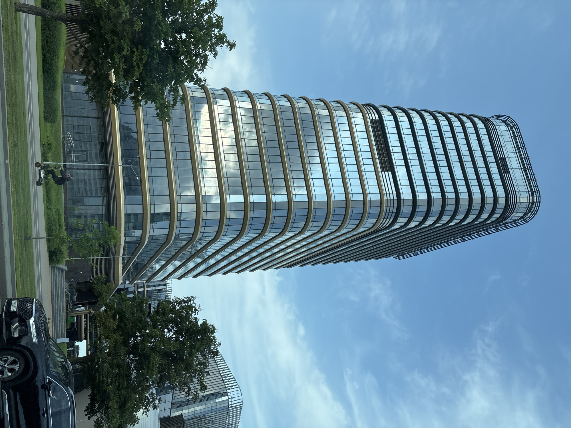 In Minsk, Belarus, a modern, multi-story office building with a curved facade and extensive glass windows stands under a partly cloudy sky. A black SUV is parked in the foreground on the right. A person is riding an electric scooter on a paved path in front of the building, passing by a street lamp and green landscaping. Another similar building is visible in the background to the right, with a large green tree in the middle ground. The overall scene depicts an urban environment.