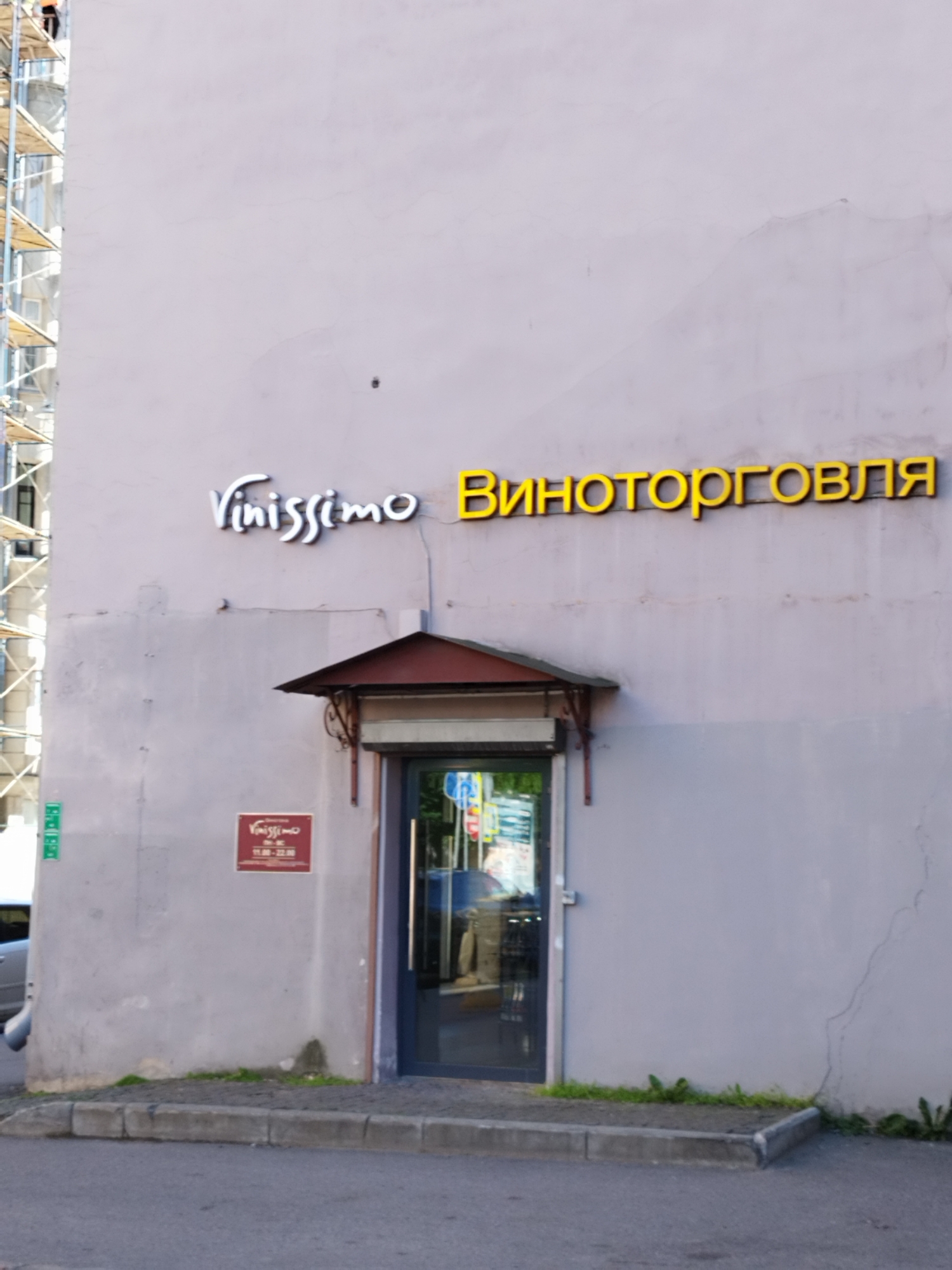This is a medium-shot, eye-level photograph of the exterior of a building, likely a storefront, on a bright, possibly sunny day. The building has a light purple-gray facade. A scaffolding structure is visible on the far left, suggesting ongoing construction or renovation.
The primary subjects are the signs and the entrance to the establishment. Above the entrance, a white, stylized sign reads "Vinissimo". To the right of this, a larger, bright yellow sign in Cyrillic script spells out "Виноторговля", which translates to "Wine Trade" or "Wine Shop".
Below the "Vinissimo" sign, and to the left of the entrance, is a small, maroon rectangular sign with "Vinissimo" also written on it, along with operating hours: "11.00 - 22.00". The entrance itself is a dark-framed glass door, partially obscured by a dark red awning. Through the glass, an interior scene is visible, hinting at the nature of the business. The street in front of the building is paved, with a concrete curb and some patches of grass and weeds growing along the building's base. A silver car is partially visible to the left. The overall impression is of a commercial establishment in a city environment.
The text visible is: "Vinissimo" and "Виноторговля" on the facade, and "Vinissimo 11.00 - 22.00" on the smaller sign. The time of day appears to be daytime, judging by the bright light and shadows. The weather seems pleasant, with clear skies. The location is implied to be Saint Petersburg, Russia, given the Cyrillic text and common architectural styles.