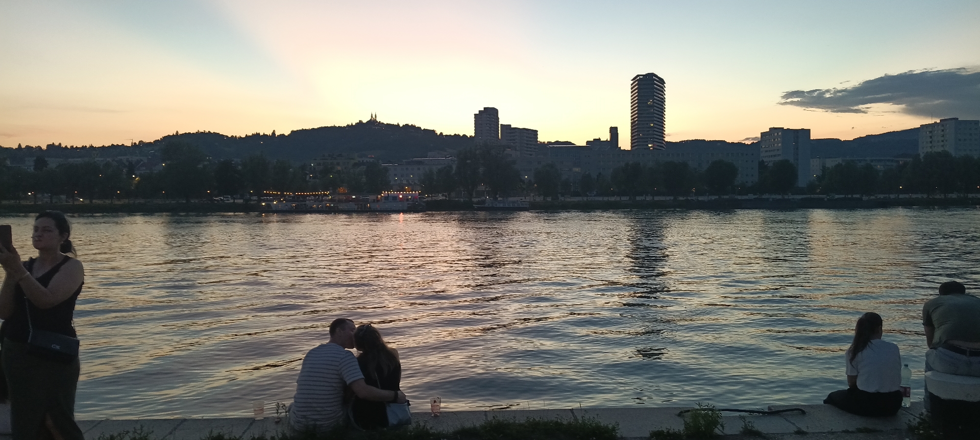 The image depicts a sunset over the Danube River in Linz, Austria. The skyline features a mix of older buildings and modern skyscrapers. In the foreground, several people are seen relaxing by the riverbank. A couple in the center is embracing and kissing, while a woman to the left is holding a phone and appearing to take a picture or video. Another person is seated on the right side of the frame, facing away from the camera. The river's surface reflects the warm colors of the sunset and the lights from the city. A few boats are visible in the distance along the opposite bank.