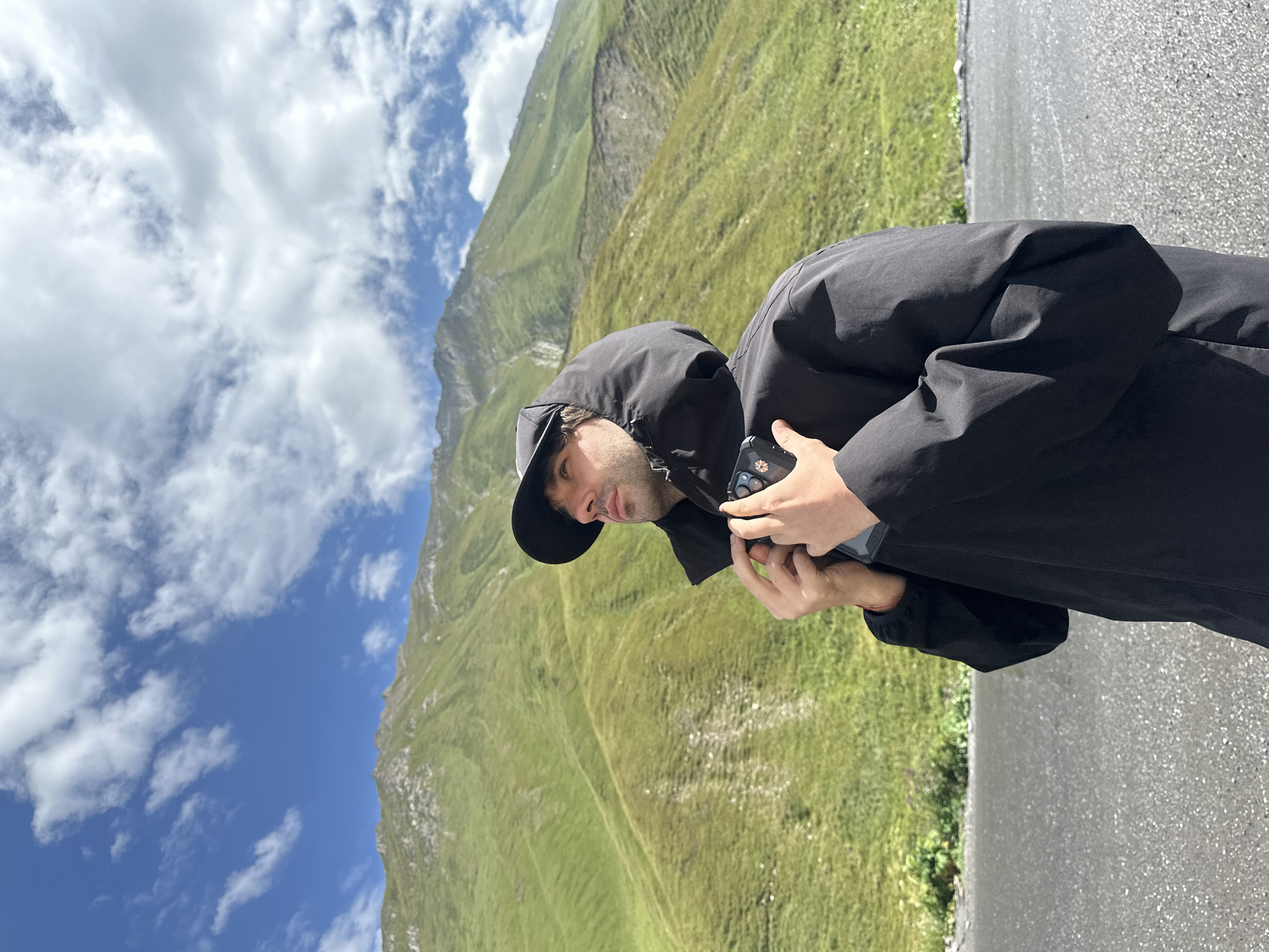 In a mountainous landscape, a man wearing a black hooded jacket and a black baseball cap is standing on a wet asphalt road. The man is looking upwards and to his right, with his hands near his chest, holding a smartphone. The background features lush green, rolling hills and mountains under a bright blue sky with scattered white clouds. The terrain appears to be a natural, outdoor environment, consistent with the Almasiani region of Georgia, known for its scenic mountain vistas.