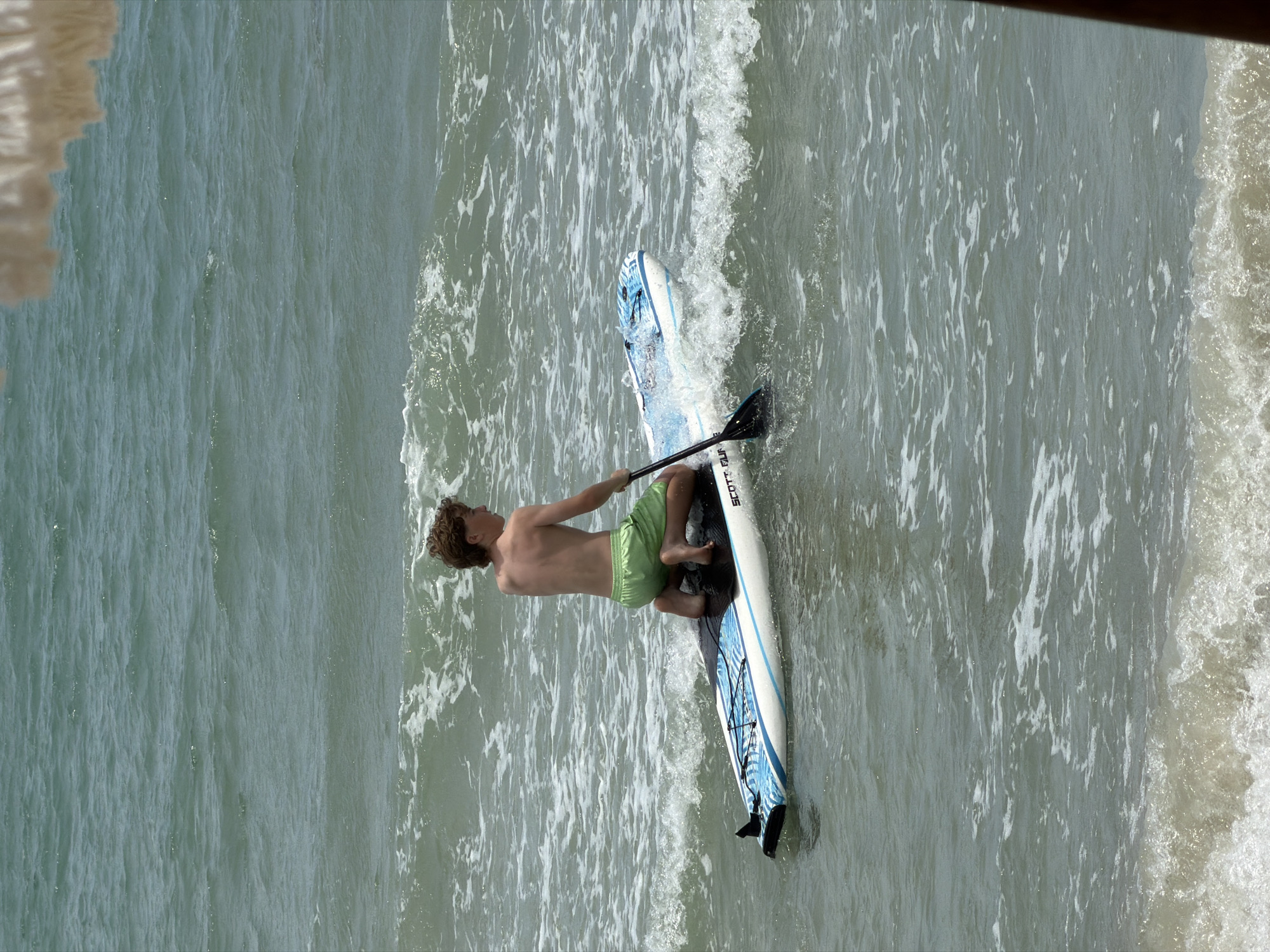 A young man with curly hair is stand-up paddleboarding in the ocean. He is shirtless, wearing light green swim trunks, and is positioned on a white and blue paddleboard. He is holding a black paddle with a blue handle, using it to propel himself through the water. The paddleboard has a blue pattern on its underside. The water is choppy, with white foam from small waves breaking around the board. The scene appears to be at a beach, likely in a tropical location given the context of Ko Samui, Thailand. The lighting suggests it is daytime. The man's body language indicates he is actively paddling and navigating the water.