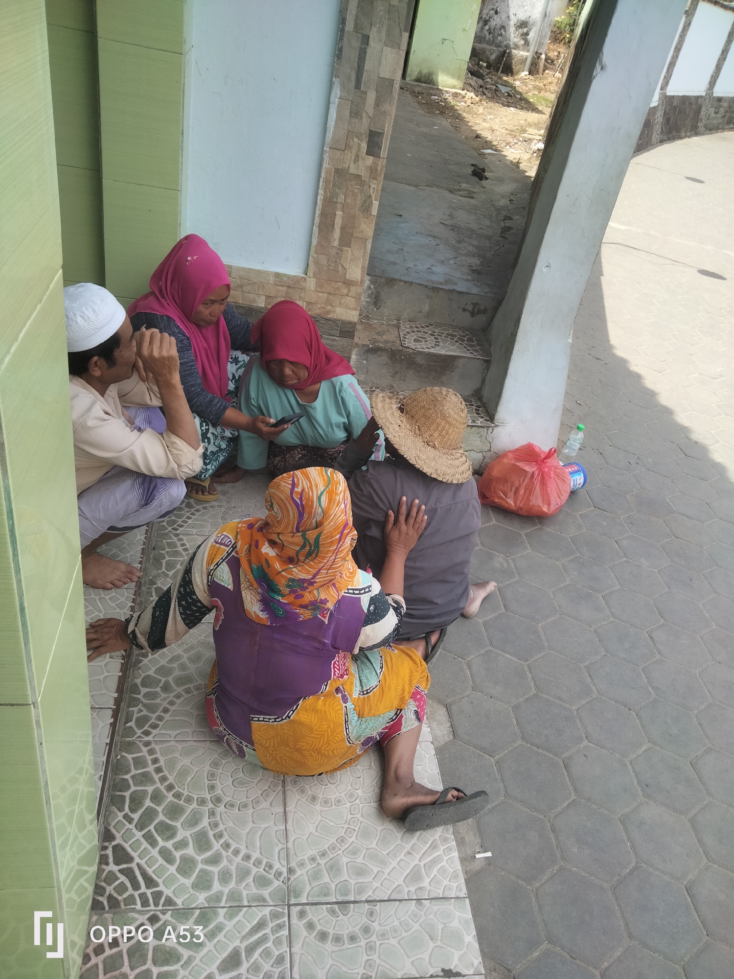 The image shows four people sitting on the tiled ground in front of a building. A man wearing a white cap and a light-colored shirt is on the left. Two women are sitting next to him, both wearing headscarves, one pink and one red. The woman in the red headscarf is holding a smartphone. Another woman, with her back to the camera, is wearing a colorful outfit and an orange and yellow headscarf. She is also wearing a straw hat. In the background, there is a red plastic bag and a bottle of water on the ground, as well as a walkway and some debris. The overall scene appears to be outdoors in a public area, possibly a side street or entrance to a building. The style of the tiles and the architecture suggest a location in Indonesia, likely in a city like Badung.