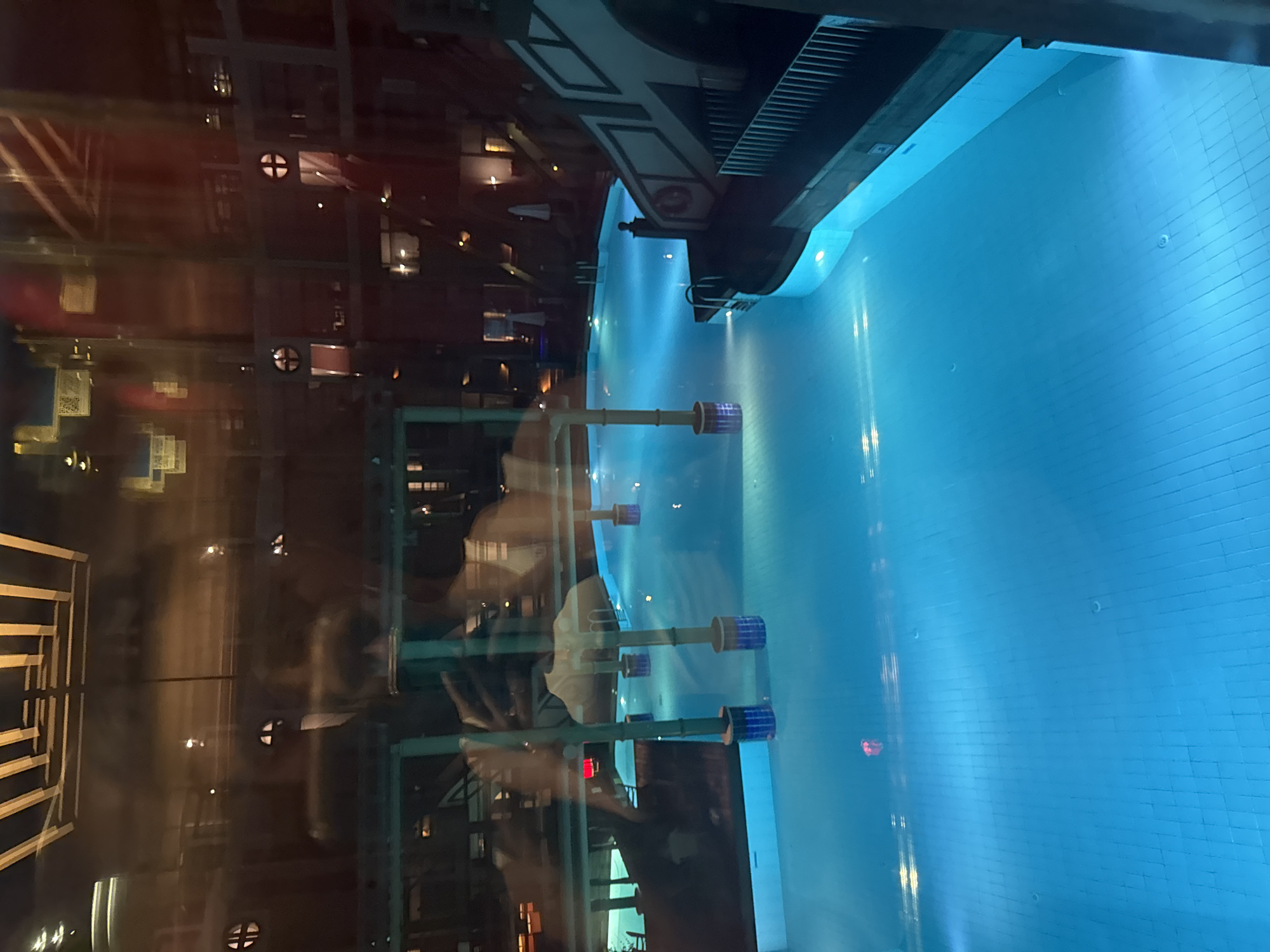 The image depicts an indoor swimming pool illuminated by blue underwater lights, creating a vibrant and inviting atmosphere. The water is clear and tranquil, with the tiled bottom of the pool visible beneath the surface. Several cylindrical structures, likely support columns or decorative elements, are submerged in the pool, their tops extending above the water.
The pool area appears to be part of a larger complex, as indicated by the reflections on the glass surface in the foreground. These reflections reveal glimpses of an indoor environment with architectural features such as wooden beams and patterned ceilings. Additionally, there are reflections of what appear to be people, suggesting that the photographer is inside a building looking out at the pool.
On the right side of the image, a section of a building's exterior is visible, characterized by a white structure with dark accents and a railing. This architectural style, along with the overall ambiance, suggests a resort or hotel setting. The presence of umbrellas in the background further reinforces this impression.
While the specific location in Aksu, Turkey, is not explicitly identifiable from the image alone, the setting is consistent with a well-appointed indoor swimming facility found in tourist destinations. The time of day is suggested to be evening or night due to the artificial lighting and the dark reflections in the glass, hinting at a serene and relaxing environment.