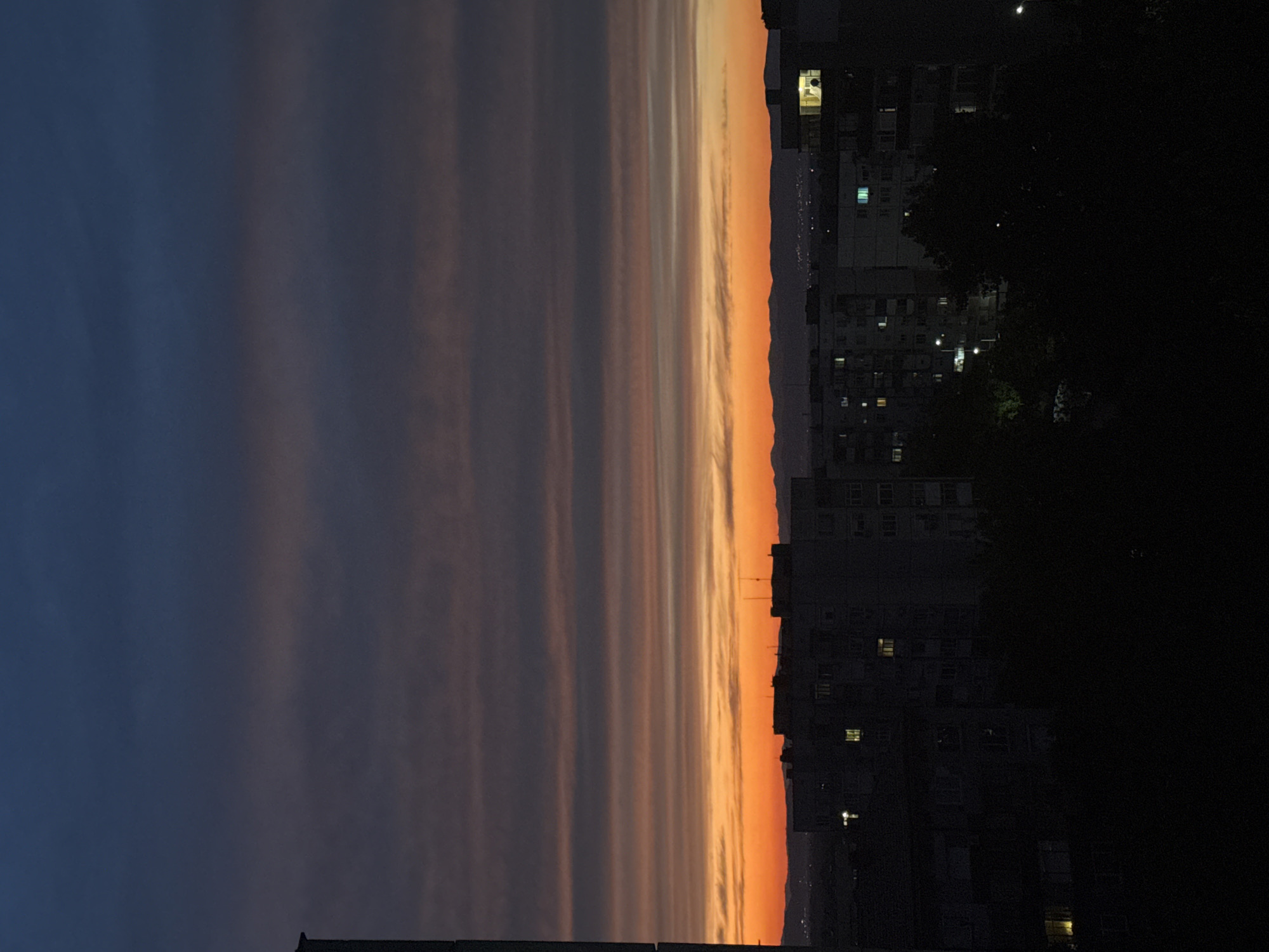 An image captures a twilight sky over an urban landscape, likely an apartment complex, in an unidentified location. The sky is a gradient of deep blue at the top, transitioning through shades of gray and purple, and culminating in a vibrant band of orange and yellow near the horizon, indicative of sunset or sunrise. Thin, horizontal clouds are visible across the sky. Below the sky, silhouetted apartment buildings with visible windows, some illuminated, fill the midground and foreground. Trees are also visible, casting dark shadows. The overall scene suggests a view from an elevated position, possibly a window or balcony, overlooking a city at dusk or dawn.