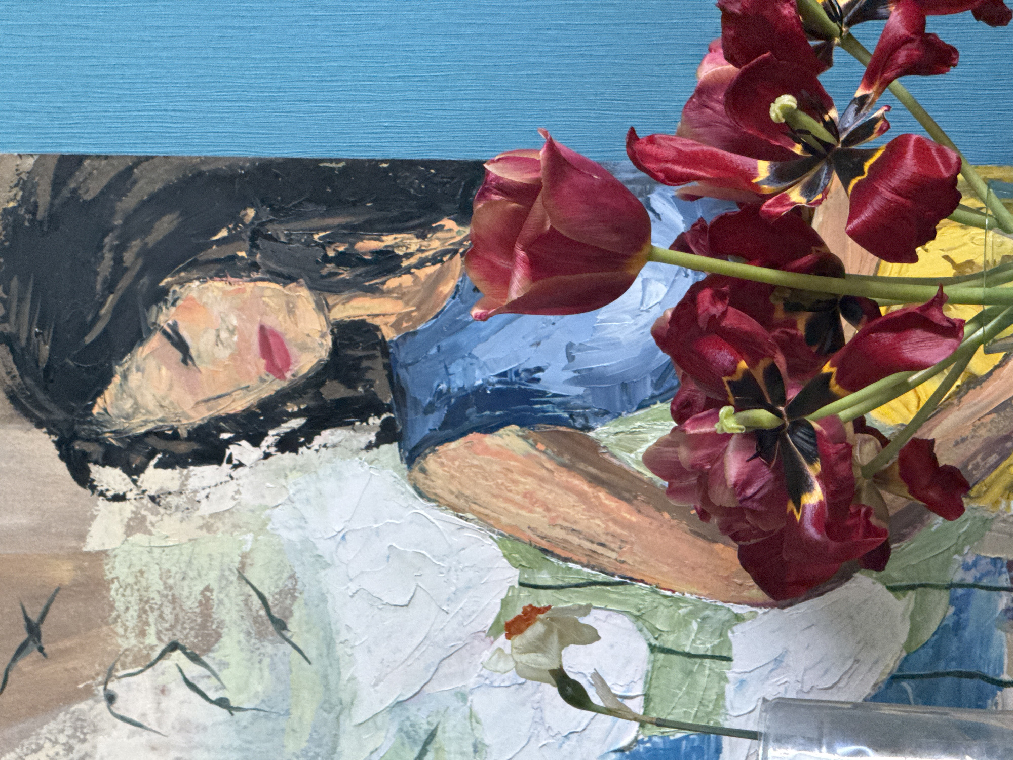 The image shows a still life with red tulips in the foreground. The tulips are at various stages of opening, with some blooms fully open, revealing black and yellow details inside. Behind the flowers, there's a painting depicting a reclining figure. The figure has dark hair, a light complexion, and red lips. The figure wears a blue garment and rests on a white surface with green and blue accents. The background above the painting is a solid blue color. A small white flower is also visible near the bottom of the frame. The location, Kazan, Russia, cannot be determined from the visual content of the image alone.
