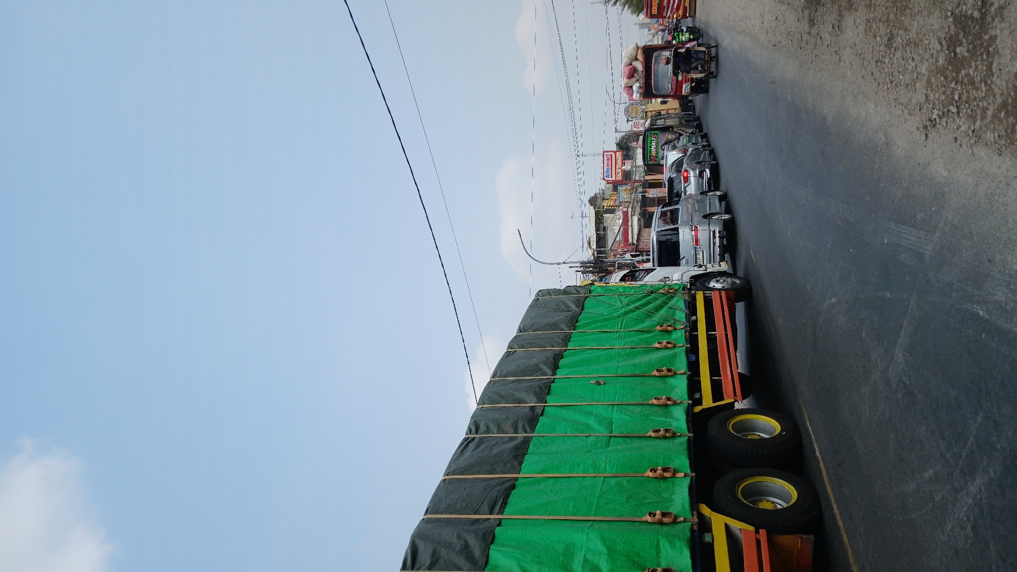 A traffic jam is captured on a road in Gombong, Indonesia, under a light blue sky with scattered clouds. The foreground is dominated by the side of a large green-covered truck. The truck has a bright yellow stripe and an orange stripe along its lower edge. The wheels of the truck are visible, with yellow hubs.
Behind the truck, a line of vehicles is visible, indicating heavy traffic. Several cars, including a silver minivan and a silver sedan, are in the queue. Further down the road, a red truck is stopped, with a person riding a motorcycle with a passenger in front of it. The red truck appears to be carrying a load of red and white bags.
Buildings line the right side of the road, with signs that are difficult to read clearly but one appears to be for "Alfamart." Overhead power lines are visible against the sky. The road surface is asphalt, with some patches of rougher material and gravel on the right shoulder. The overall scene depicts a congested urban or suburban roadway.