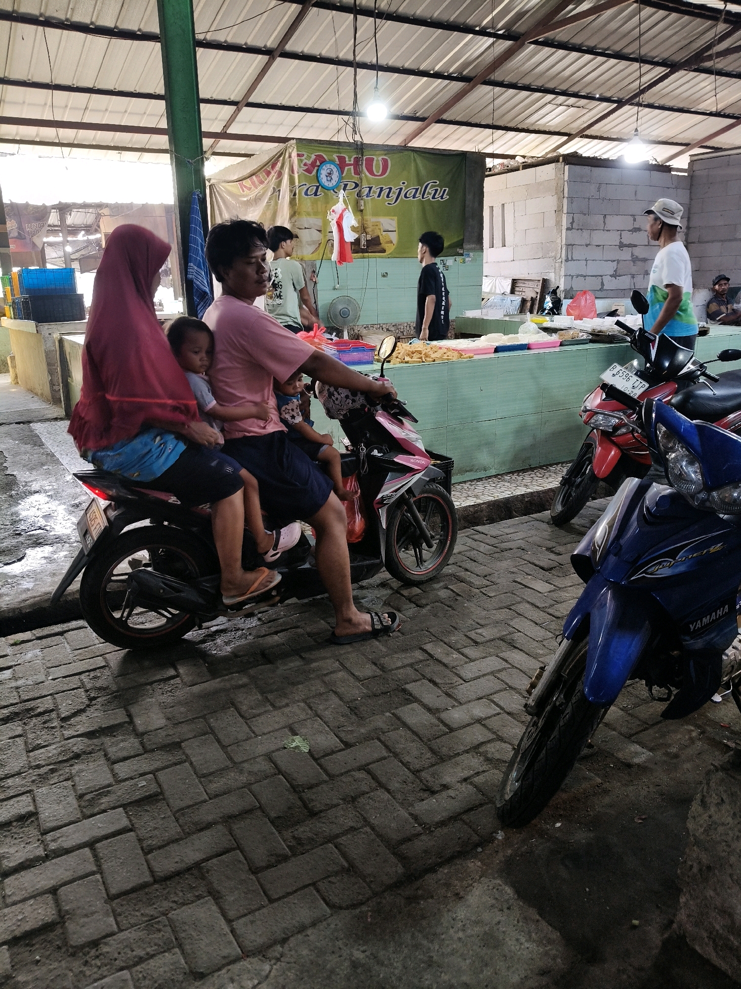 In Setiamekar, Indonesia, at a market, a family of four is seen on a motorbike. A woman in a pink hijab and a blue and white outfit is on the back, with two young children. One child is in front of her, and another is behind her. The driver, a man in a pink shirt and dark shorts, is steering the motorbike. The market appears to be open-air with a metal roof supported by green beams. Stalls with food and goods are visible in the background, along with other motorbikes and people. The ground is paved with dark bricks.