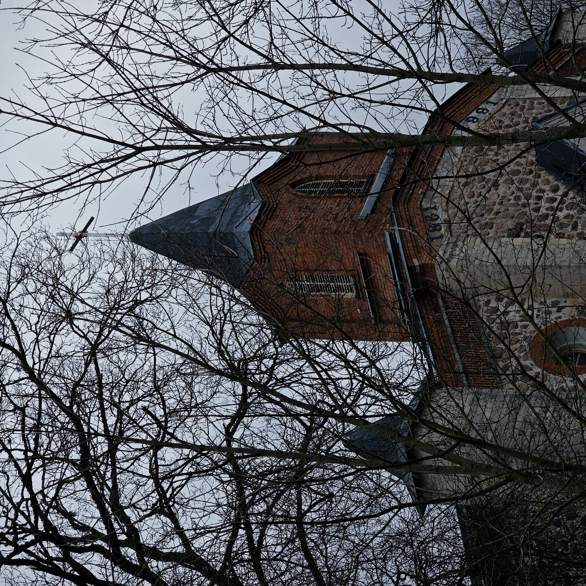 A low-angle view of a brick and stone building topped with a peaked roof and cross is obscured by bare branches. The building, identifiable as a church in Gubanitsi, Russia, is partially made of red brick and partially of rough-hewn stones. The number "1881" is visible on one section. Branches fill the foreground and surround the building, hinting at a possibly rural or park-like setting. The sky is overcast. No people or activities are visible.