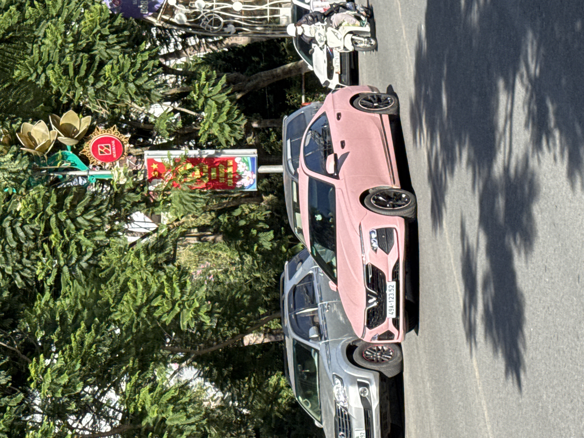 The image captures a street scene in Da Lat, Vietnam, on a sunny day. The foreground features a pink sedan with a Vietnamese license plate reading "49A-123.52." To the left of the pink car, a silver SUV with a Vietnamese license plate is parked. In the background, trees provide shade, and a few street decorations are visible, including a red banner with Vietnamese text and graphics, and a circular emblem with the AgriBank logo. A person on a white scooter, wearing a face mask and a jacket, is moving away from the pink car towards the right side of the frame. Another white car is parked behind the scooter. The asphalt road is visible in the foreground, with shadows cast by trees.