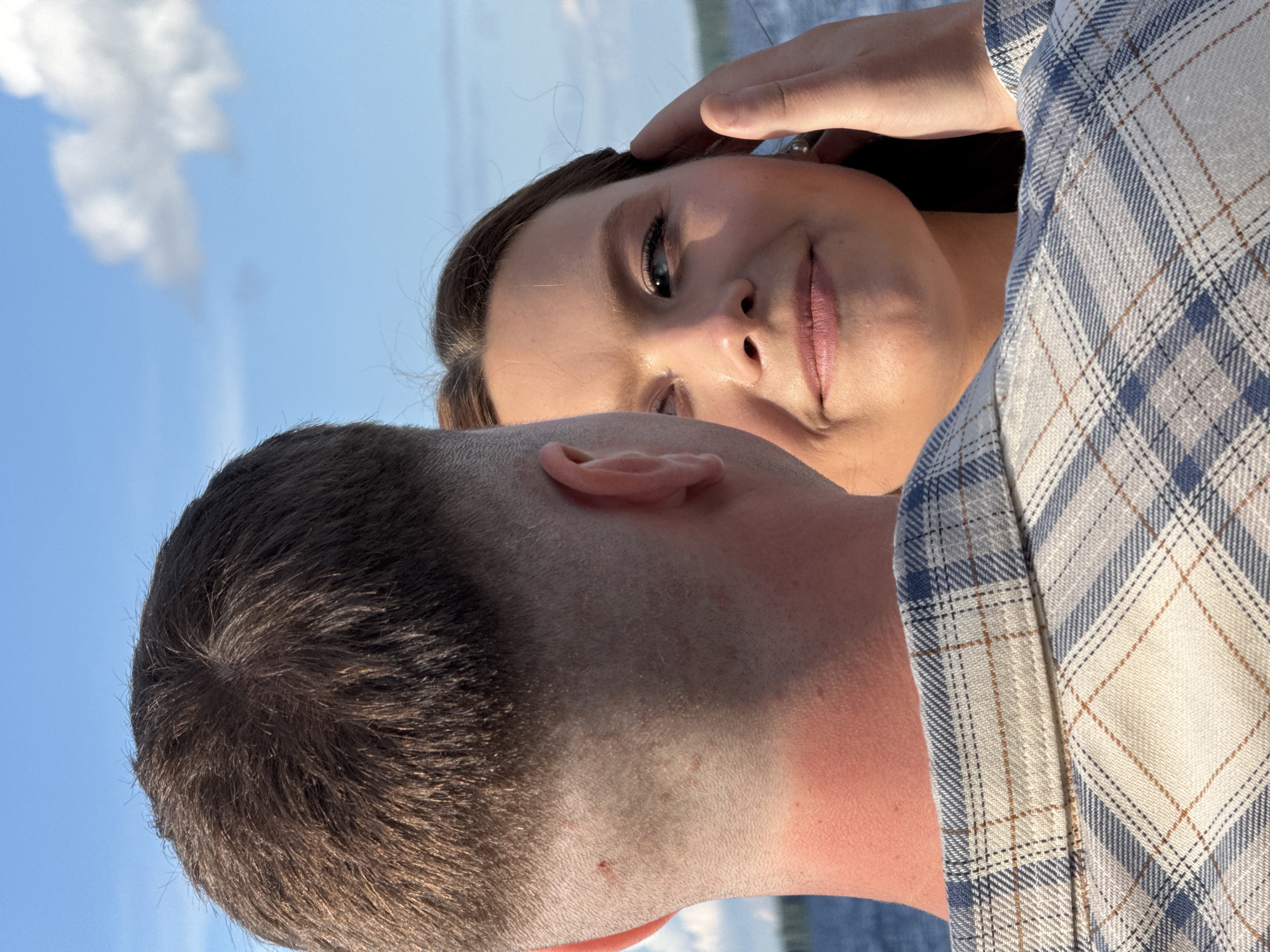 The image shows a close-up of two individuals in an outdoor setting, likely near water, given the presence of blue sky and what appears to be a body of water in the background. The foreground features the back of a man's head with a short haircut and light skin. His right ear is visible. He is wearing a light-colored plaid shirt with blue and brown patterns. Partially obscured by the man is a woman with brown hair. Her left hand is gently touching her face. The facial expression is neutral. The photo appears to have been taken during the daytime, as there is ample sunlight illuminating the subjects. There is no specific evidence to suggest the location is Chaikovsky, Russia, and that cannot be conclusively stated without additional information.