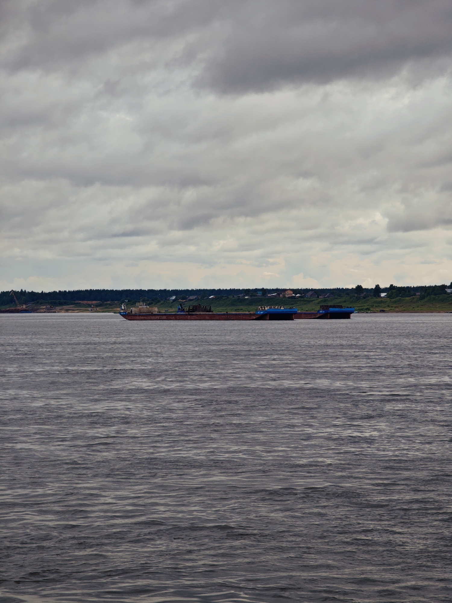 In Zotino, Russia, a wide, dark river flows under a cloudy, overcast sky. Two barges, one larger and rust-colored, the other a lighter blue, are moored in the river. The barges are laden with cargo. In the background, a lush green forest lines the riverbank, interspersed with a few small houses, indicating a sparsely populated settlement. The overall scene suggests a tranquil, perhaps somber, atmosphere common in northern climates.