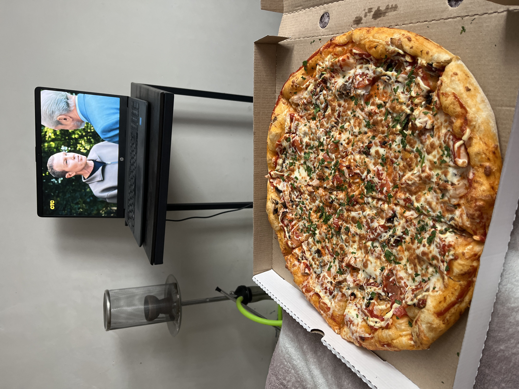 A pizza in an open cardboard box sits on a surface beside a hookah and a laptop displaying two men on its screen. The pizza is round with a thick crust and is covered in melted cheese, tomato slices, and sprinkled with parsley. The hookah is silver with a mesh-covered bowl and a green hose. The laptop, showing two men in professional attire, is positioned on a black stand. The background consists of a plain, light-colored wall. The scene is indoors. It is not possible to determine if this image was taken in Verkhnyaya Pyshma, Russia from the provided information.
