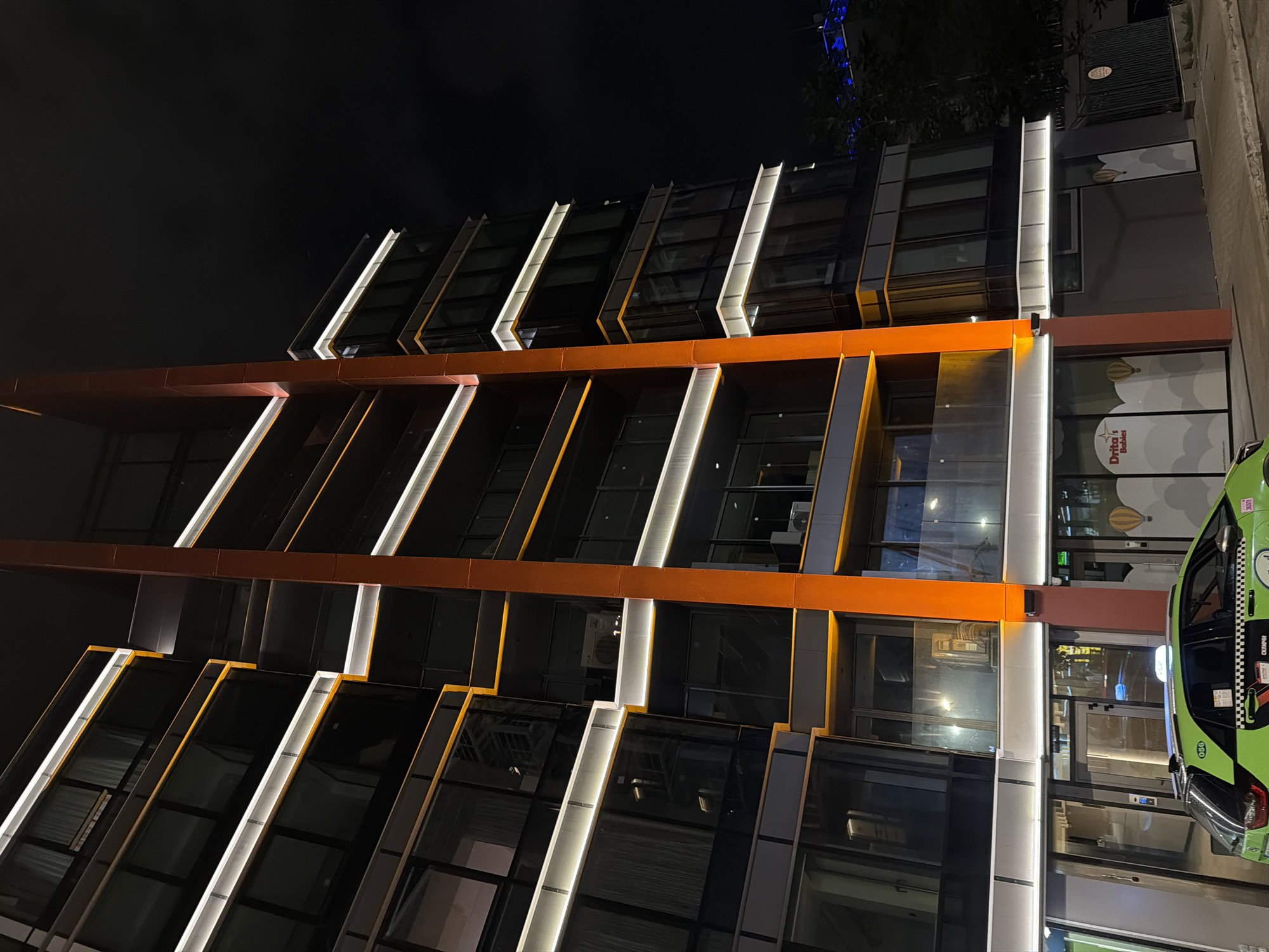 The image captures a modern building facade at night in Sofia, Bulgaria. The building features a sleek, angular design dominated by extensive glass panels set within a framework of dark gray and silver elements. A prominent orange horizontal band cuts across the middle section of the facade. Bright, cool-toned linear LED lights are integrated vertically into the building's structure, creating striking illuminated lines that contrast sharply with the dark glass windows and the nighttime sky. These lights emphasize the geometric patterns of the architecture.
The sky is dark, indicating nighttime, and the artificial lighting from the building and street elements is the primary illumination source. Reflections are visible on the glass surfaces of the building.
On the lower right side of the building, a green taxi is partially visible, showing its front-right side, including a headlight, part of the windshield, and a side mirror. The word "OK" is visible on the front of the taxi.
Further up on the right, on the building's lower section, a storefront sign is displayed. The sign clearly reads "Drita's Babies" and includes a website address, "www.dritasbabies.com," below it. The sign also features hot air balloon illustrations as part of its design.
The scene is static, depicting an urban environment without any explicit human activity, focusing on the architectural details and nighttime illumination of the structure.