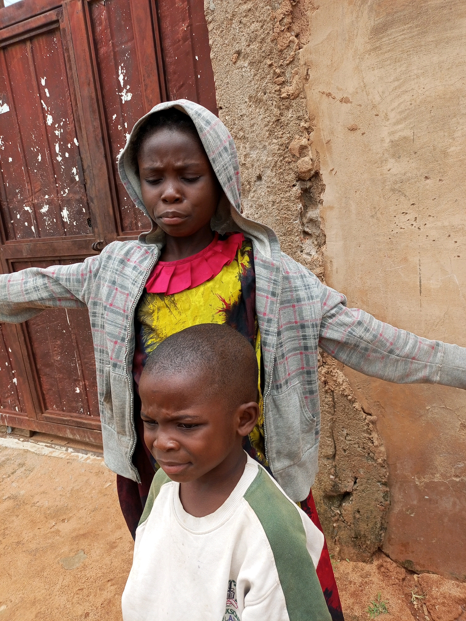 Two children are standing in front of a reddish-brown, weathered door and a textured, ochre-colored wall in what appears to be a village setting in Tempure, Nigeria. The child in the foreground, a young boy, wears a white sweatshirt with green accents. He is looking down and to his right with a concerned expression. Behind him, a slightly older child, presumably a girl, is wearing a grey plaid hooded jacket over a brightly colored yellow and red dress. Her eyes are closed, and her head is tilted down. Her arms are outstretched to her sides. The ground is composed of sandy soil with some sparse vegetation.